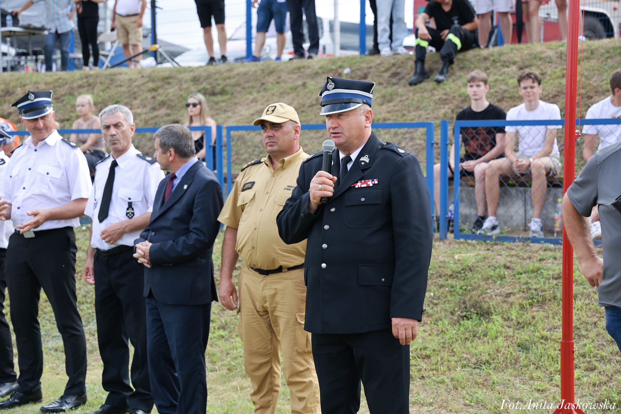 Mężczyzna w czarnym mundurze z czapką trzyma mikrofon, obok inni mężczyźni w mundurach i garniturze, w tle siedzą młodzi ludzie za niebieskim płotem.
