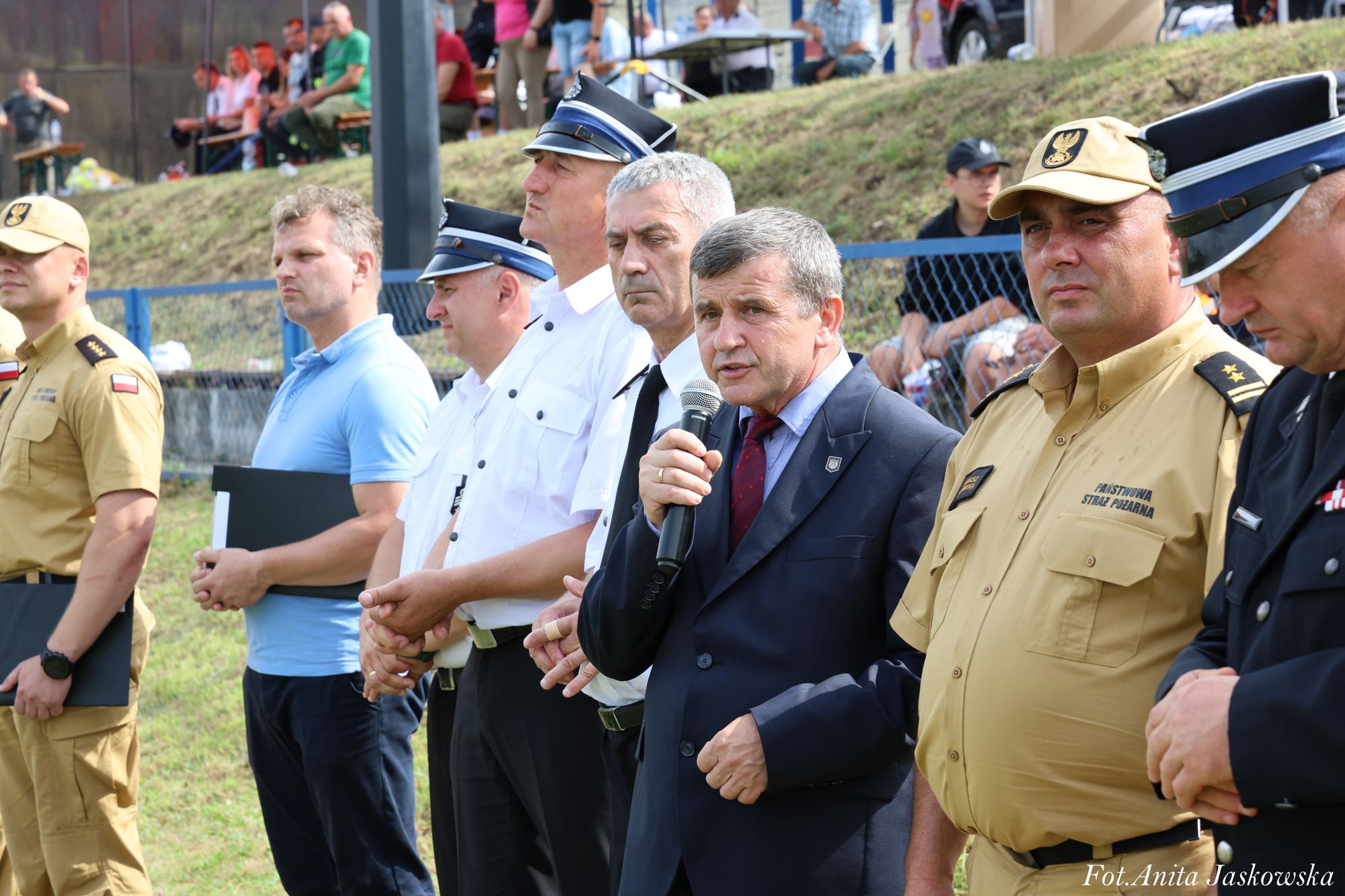 Grupa mężczyzn w różnych mundurach i garniturze stoi w rzędzie na zewnątrz, jeden z nich mówi do mikrofonu, w tle widoczni ludzie na trawie i ławkach.