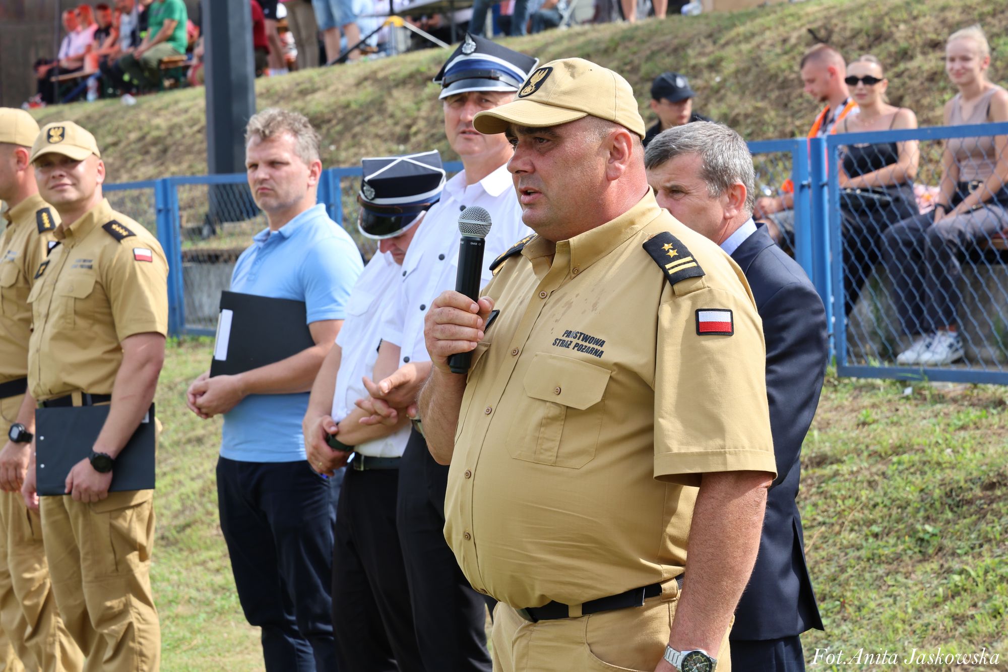 Mężczyzna w beżowym mundurze Państwowej Straży Pożarnej z mikrofonem, obok inni mężczyźni w mundurach i ubraniach cywilnych, w tle ogrodzenie i osoby siedzące na trawie.