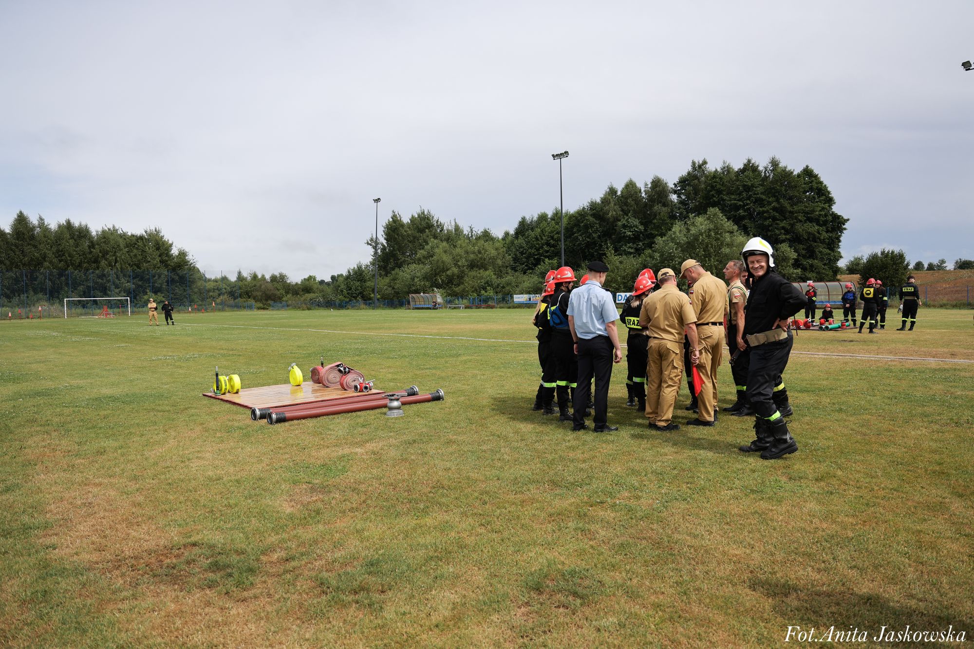 Grupa strażaków w hełmach i mundurach stoi na zielonym boisku, obok leżą zwinięte węże strażackie i drewniana podkładka, w tle drzewa i szare niebo.