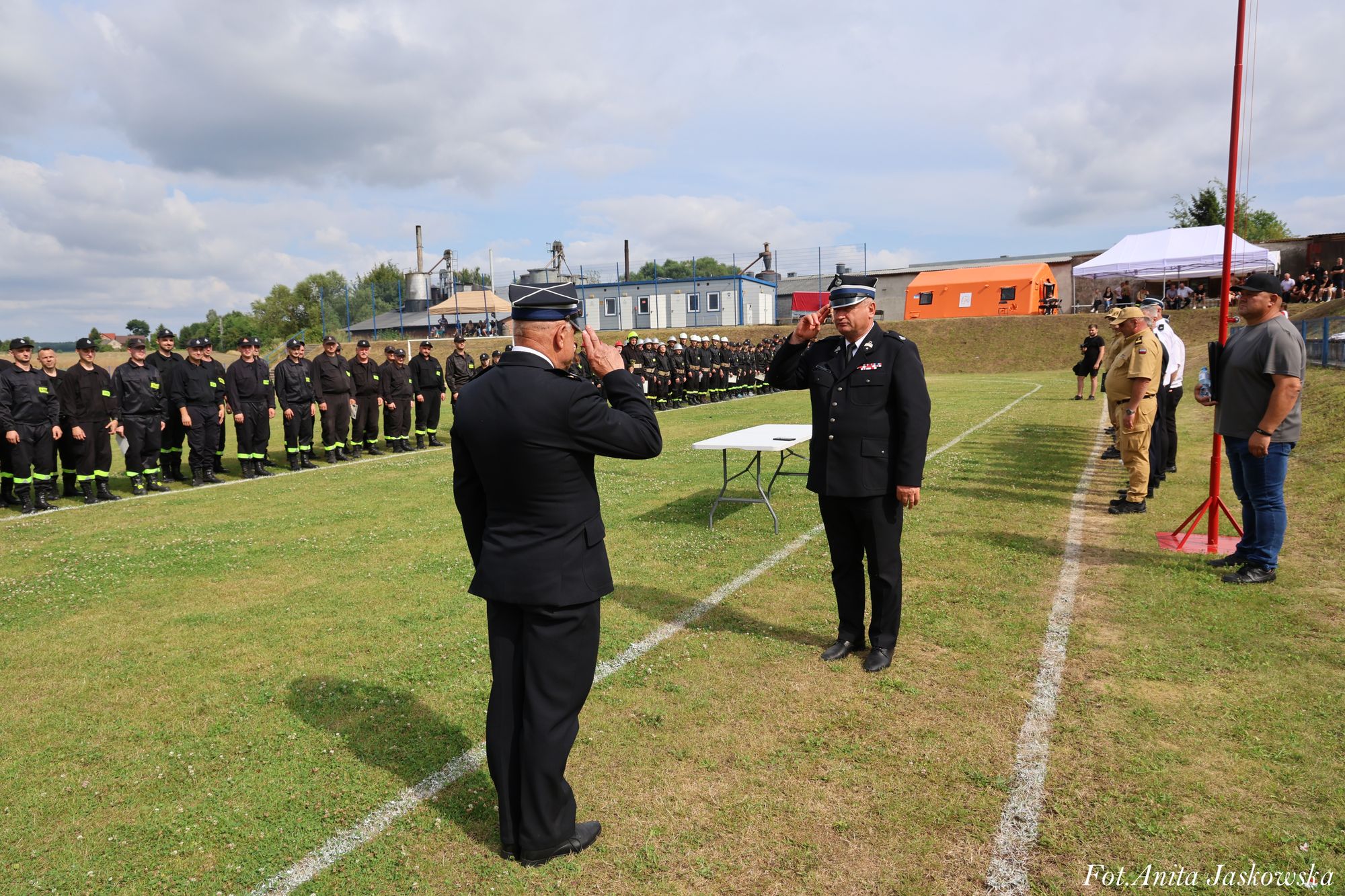 Dwóch mężczyzn w mundurach pozdrawia się na trawie, w tle rząd osób w czarnych mundurach i kilka osób stojących po prawej stronie obok stołu.