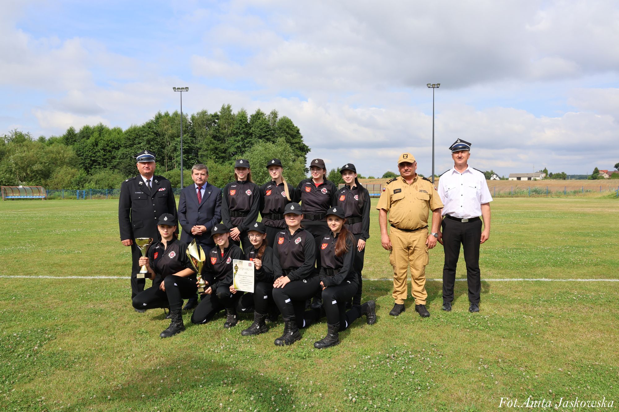 Grupa osób na trawie, większość w czarnych mundurach z czapkami, trzy osoby stojące w różnych mundurach, dwie dziewczyny trzymają puchary i dyplom.