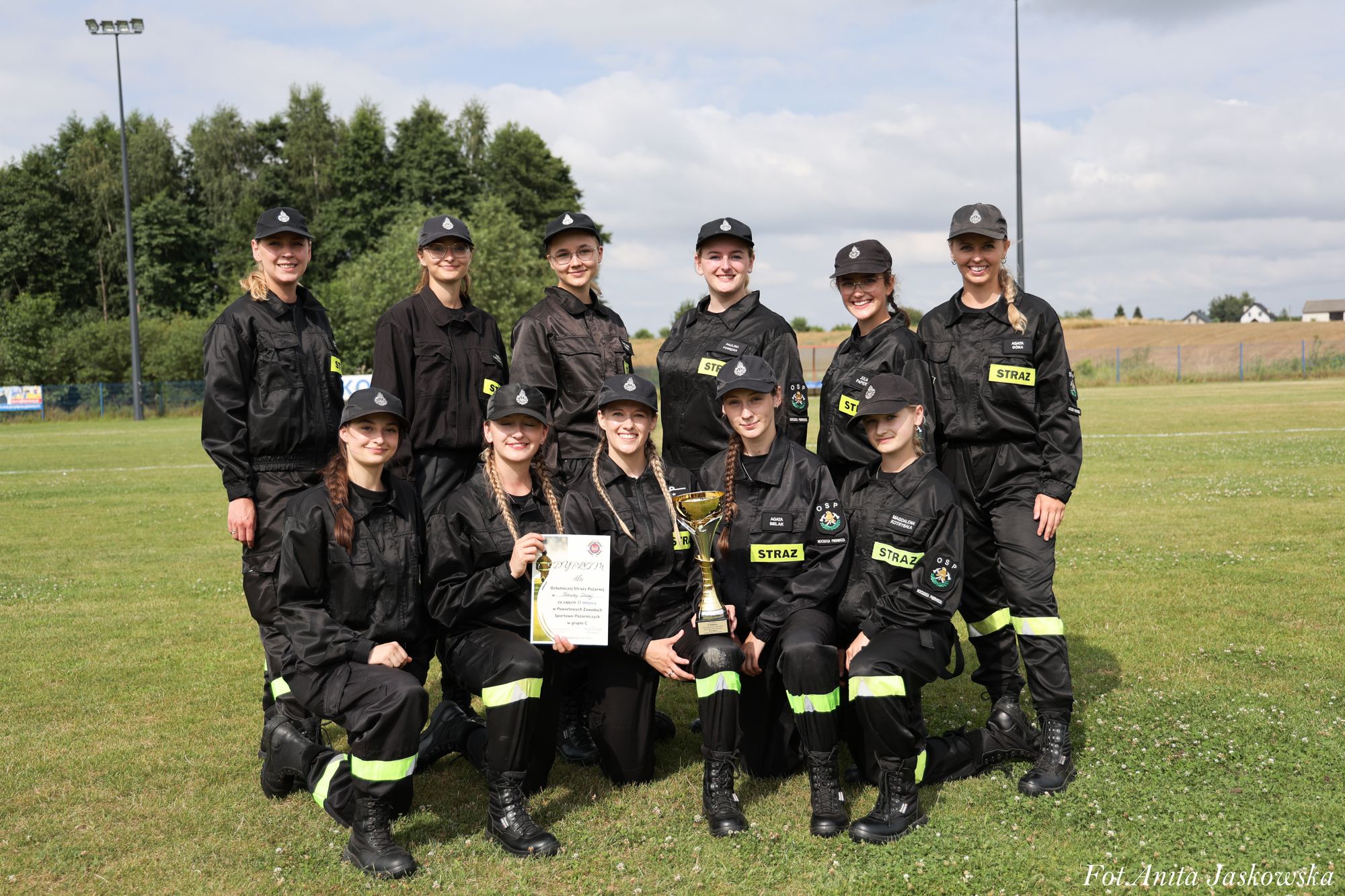 Dziewięć kobiet w czarnych mundurach strażackich, trzy klęczą, sześć stoi, trzymają puchar i dyplom na zielonym boisku, w tle drzewa i niebo z chmurami.