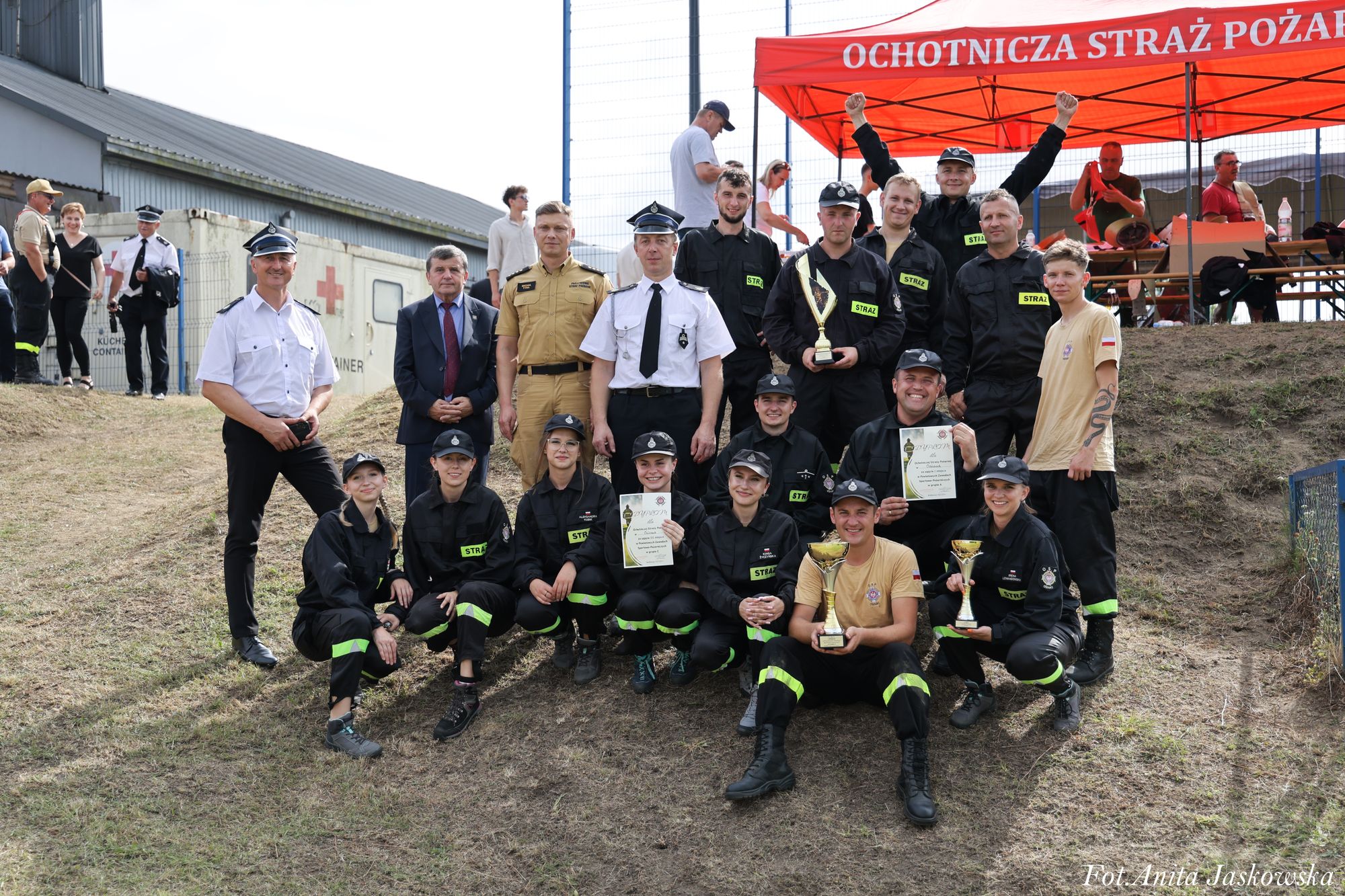 Grupa osób w mundurach strażackich i garniturowych na trawie, niektórzy trzymają puchary i dyplomy, w tle czerwony namiot z napisem "Ochotnicza Straż Pożarna".