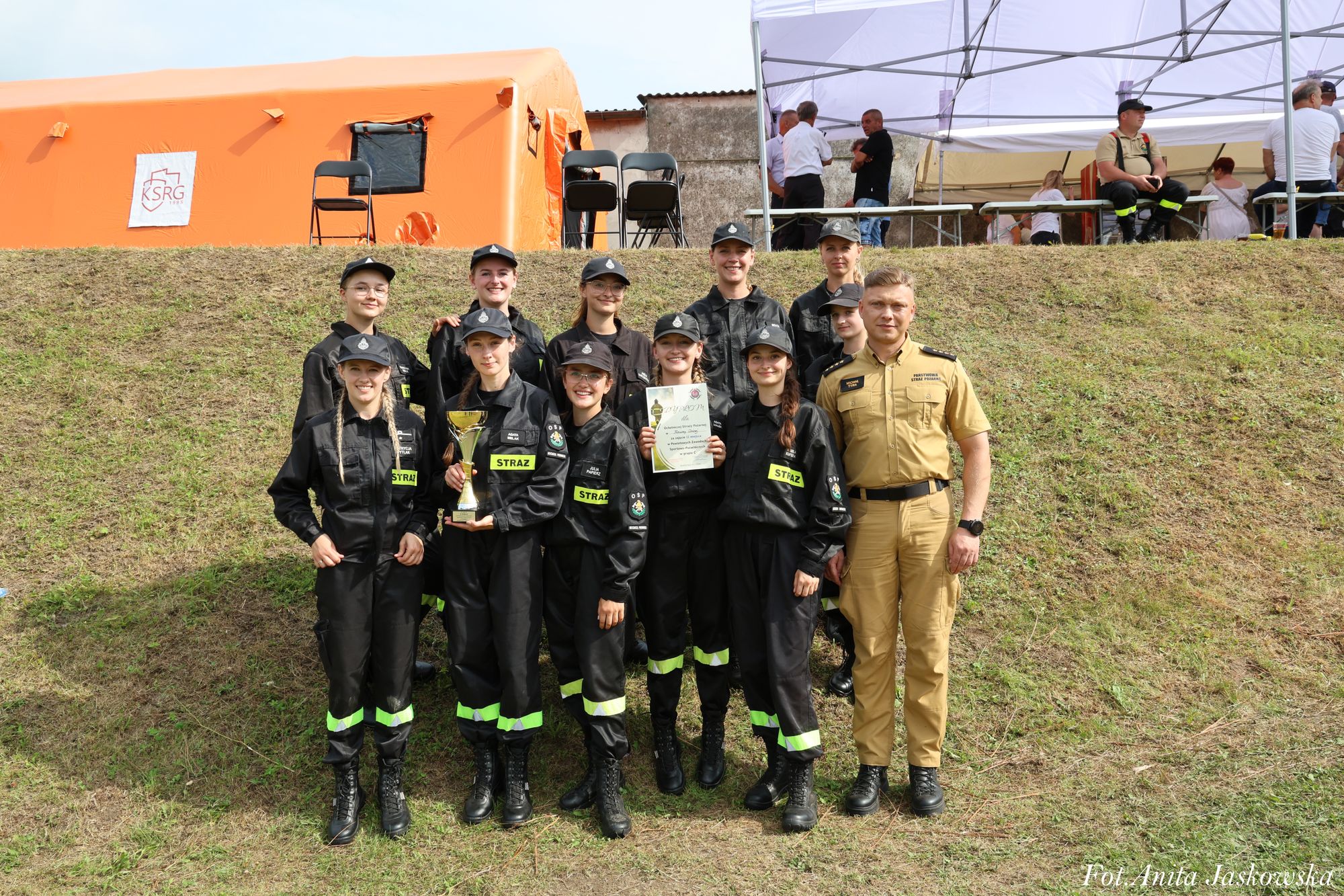 Grupa młodych strażaków w czarnych mundurach z odblaskowymi pasami, trzymająca puchar i dyplom, obok mężczyzna w beżowym mundurze, w tle pomarańczowy namiot i ludzie pod białym daszkiem.