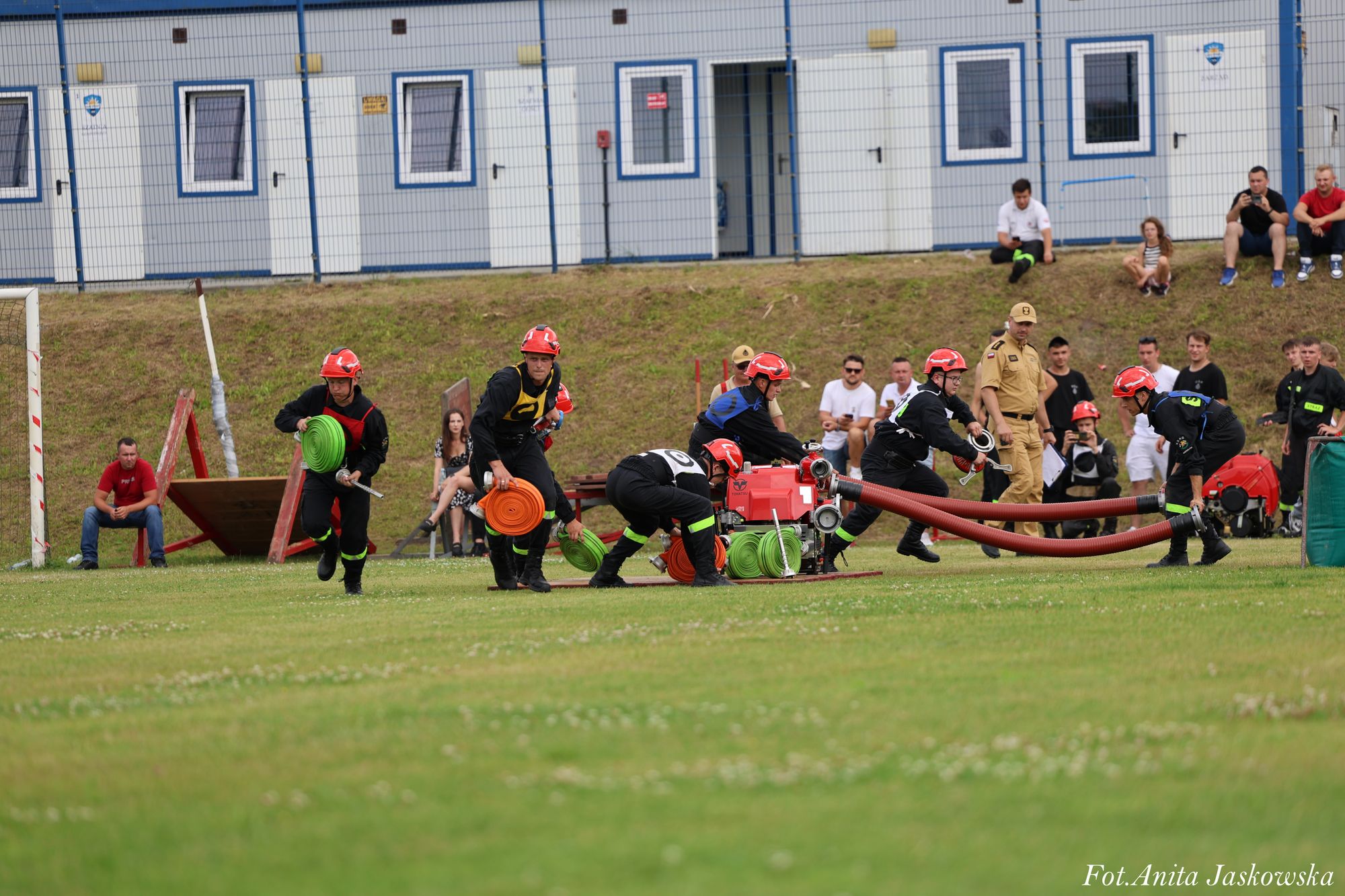 Grupa strażaków w czarnych mundurach i czerwonych hełmach podczas ćwiczeń na zielonym boisku, w tle widoczne kontenery i ludzie obserwujący zawody.