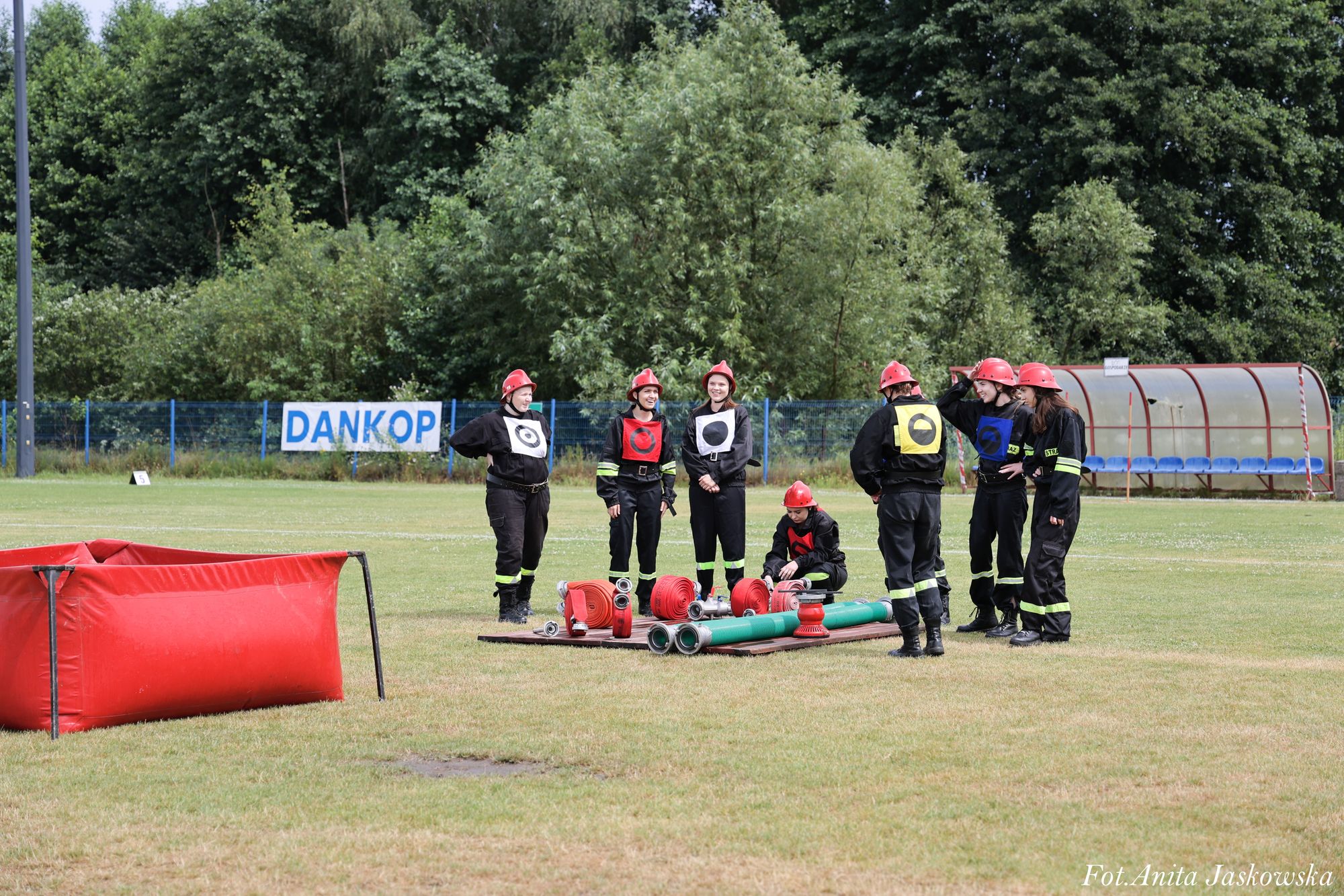 Siedem osób w czarnych kombinezonach i czerwonych hełmach stoi i kuca na zielonym boisku obok zrolowanych węży strażackich i czerwonego zbiornika na wodę, w tle drzewa i niebieska siatka.