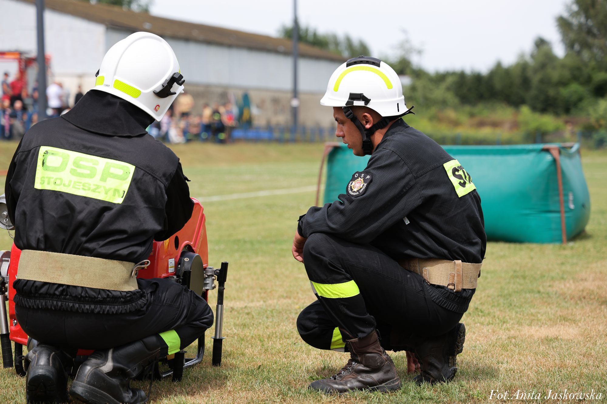Dwóch strażaków w białych hełmach i czarnych mundurach z żółtymi oznaczeniami kuca na zielonej trawie przy sprzęcie pożarniczym. W tle rozmyte osoby i budynki.