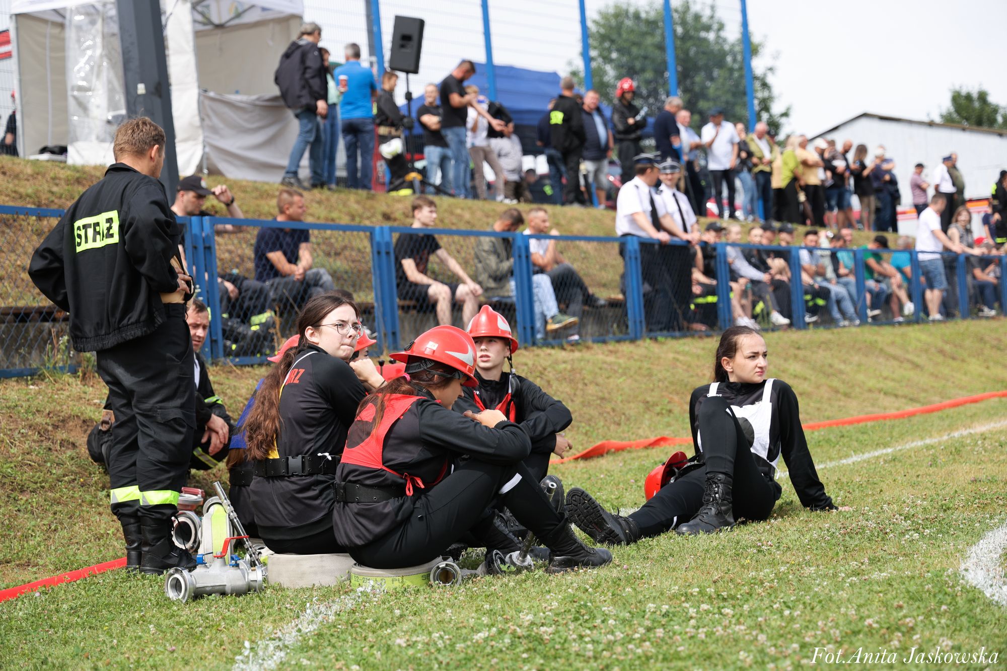 Grupa osób w czarnych ubraniach z czerwonymi hełmami siedzi i stoi na trawie przy niebieskich barierkach, w tle tłum ludzi na podwyższeniu.