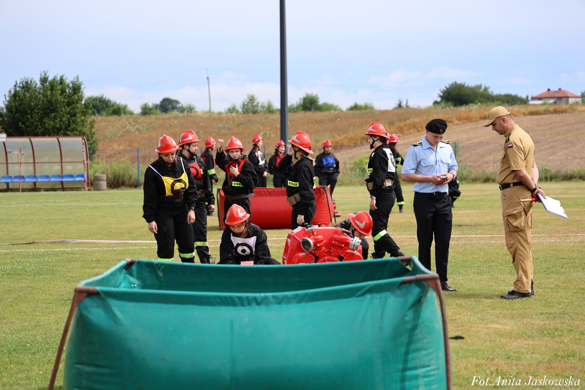 Grupa osób w czarnych uniformach i czerwonych hełmach stoi na zielonym boisku, dwóch mężczyzn w mundurach obserwuje zawody strażackie.