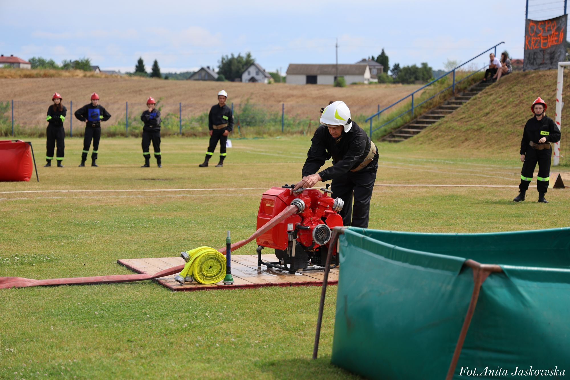Strażak w białym hełmie obsługujący czerwoną pompę wodną na trawie, w tle pięć osób w czarnych uniformach i czerwonych hełmach, zielony zbiornik na wodę.