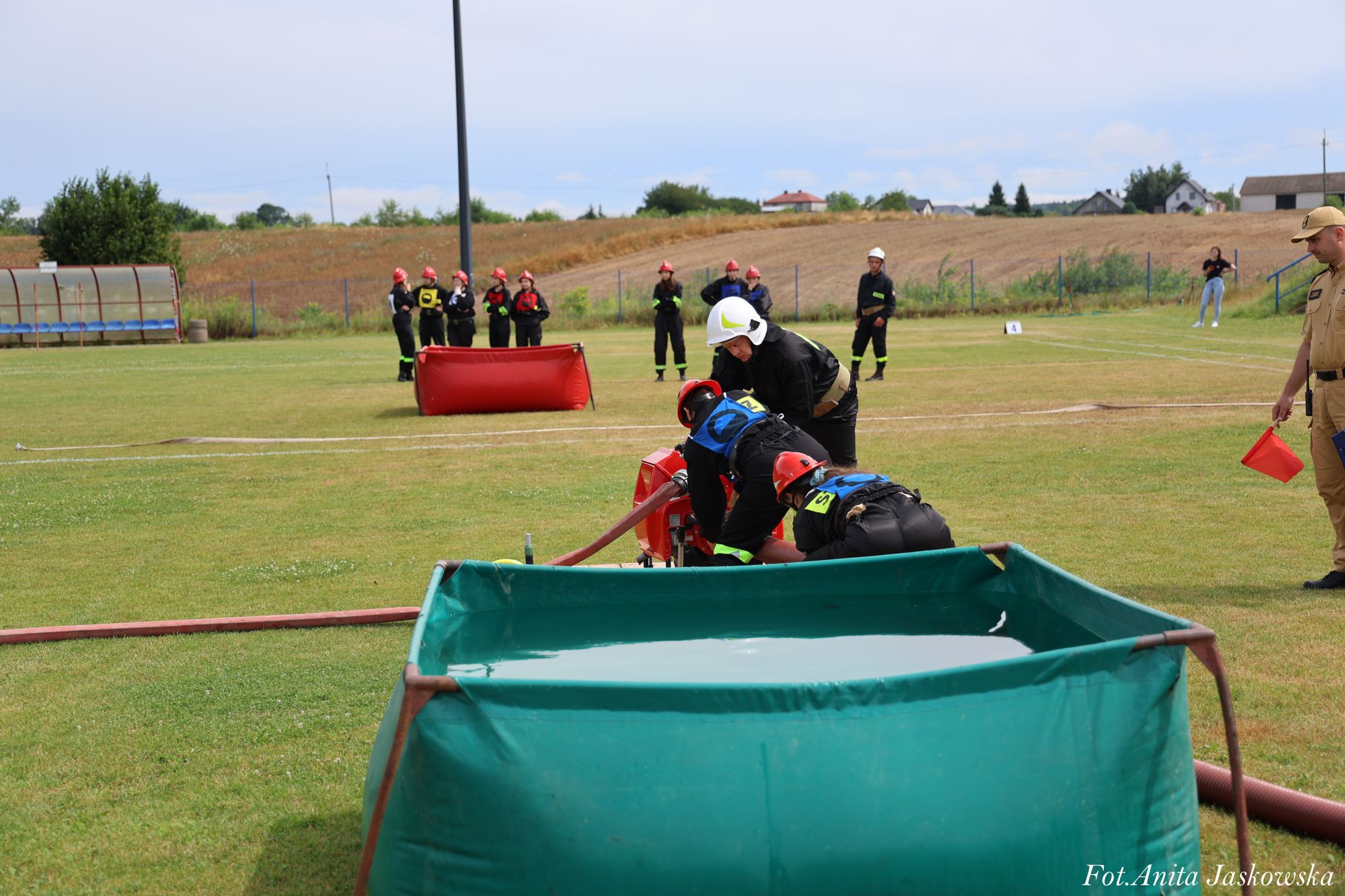 Trzech strażaków w hełmach przy pompie wodnej na zielonym polu, w tle inni strażacy i osoba z czerwoną flagą, niebo częściowo zachmurzone.