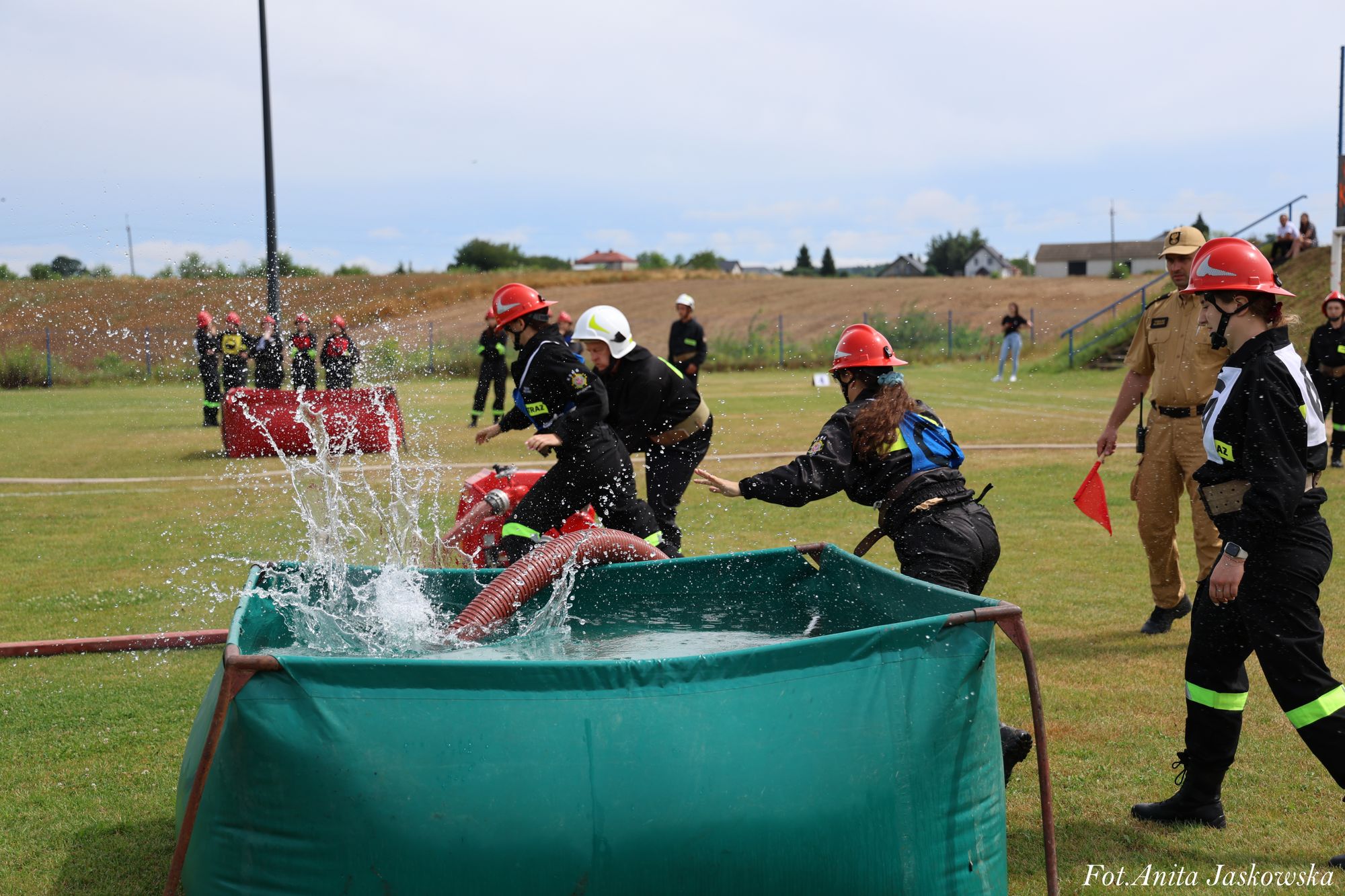 Zielony zbiornik z wodą i rozpryskującą się wodą, osoby w czarnych ubraniach strażackich i czerwonych hełmach, pole i budynki w tle.