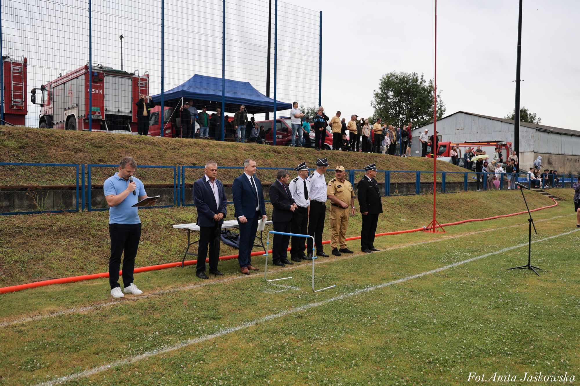 Grupa mężczyzn w mundurach i garniturach stoi na trawie, z tyłu ogrodzenie, ludzie na wzniesieniu, w tle wozy strażackie i niebieski namiot.