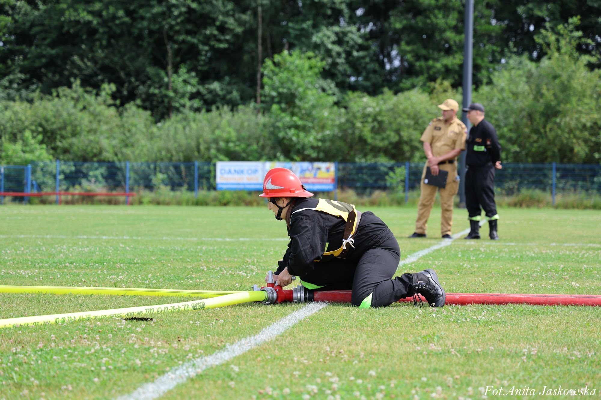 Osoba w czarnym ubraniu ochronnym i czerwonym hełmie klęczy na zielonej trawie, podłączając żółty wąż do czerwonego sprzętu, w tle dwie osoby w mundurach na boisku.