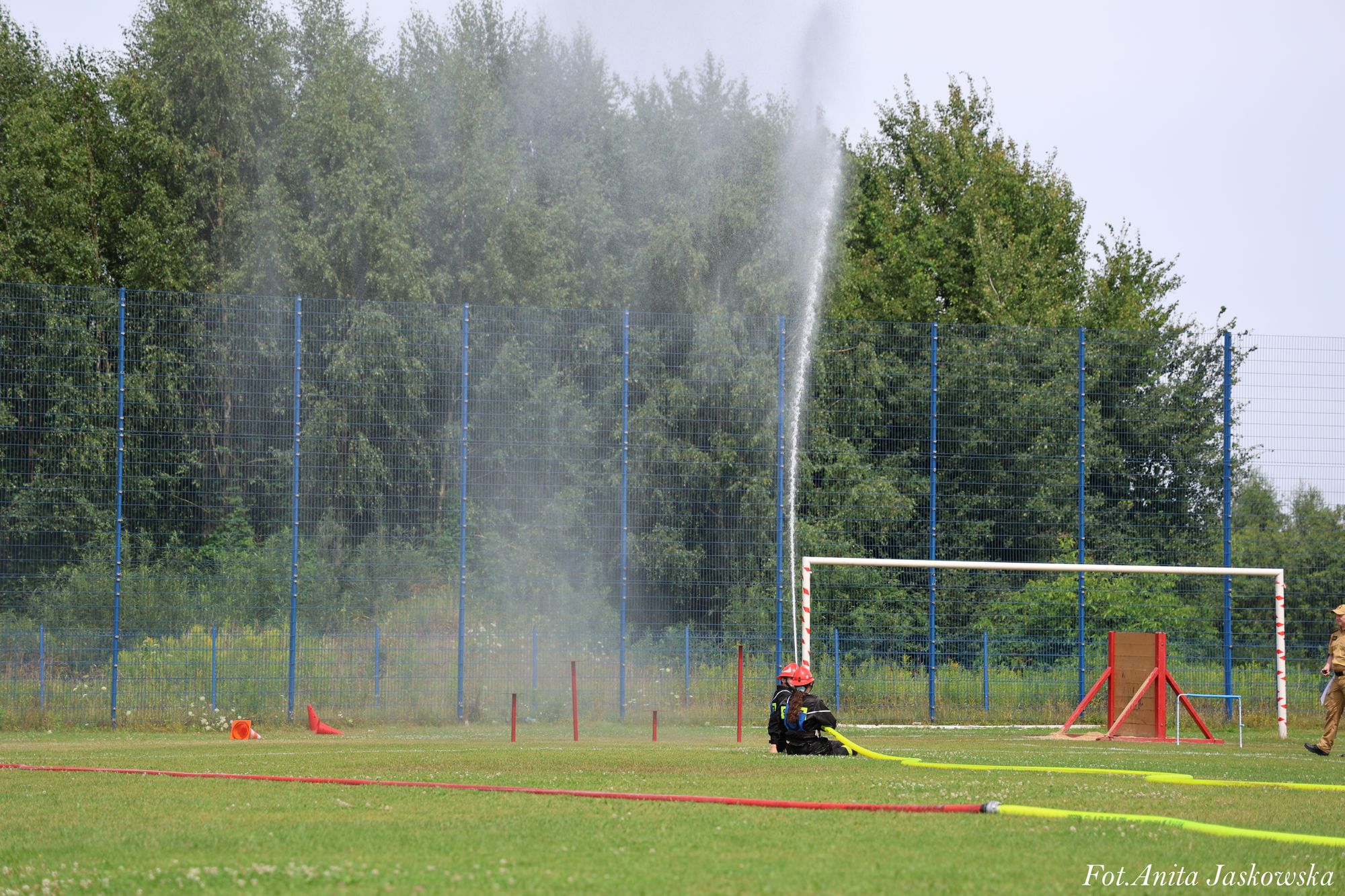 Dwoje strażaków w ciemnych ubraniach i czerwonych hełmach na zielonym boisku trzymający żółty wąż z którego leci strumień wody, w tle bramka i niebieskie ogrodzenie na tle drzew.