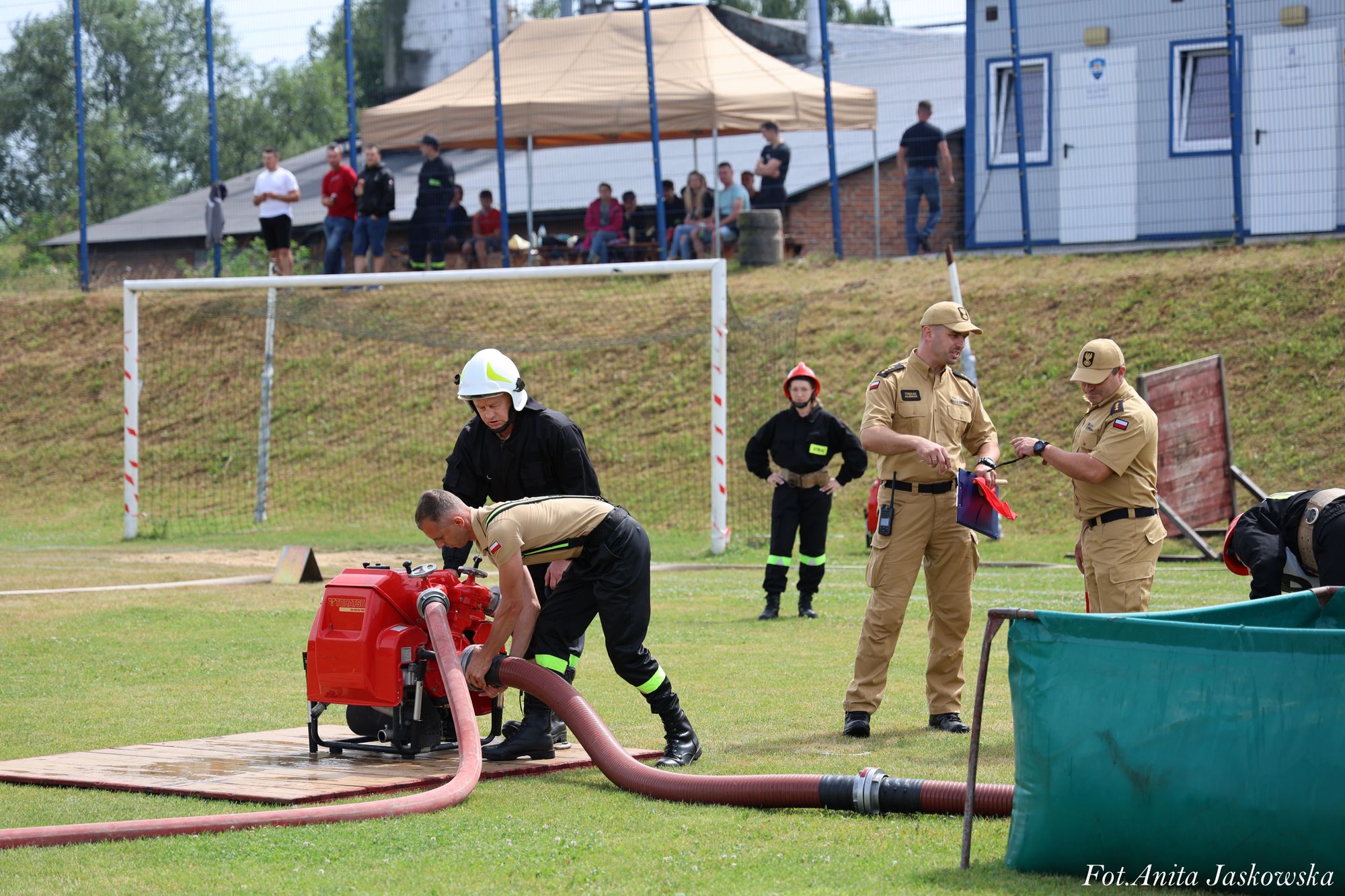Strażacy w beżowych i czarnych mundurach przy czerwonej pompie wodnej i zielonym zbiorniku na trawie, w tle ludzie pod namiotem i budynki, bramka piłkarska.
