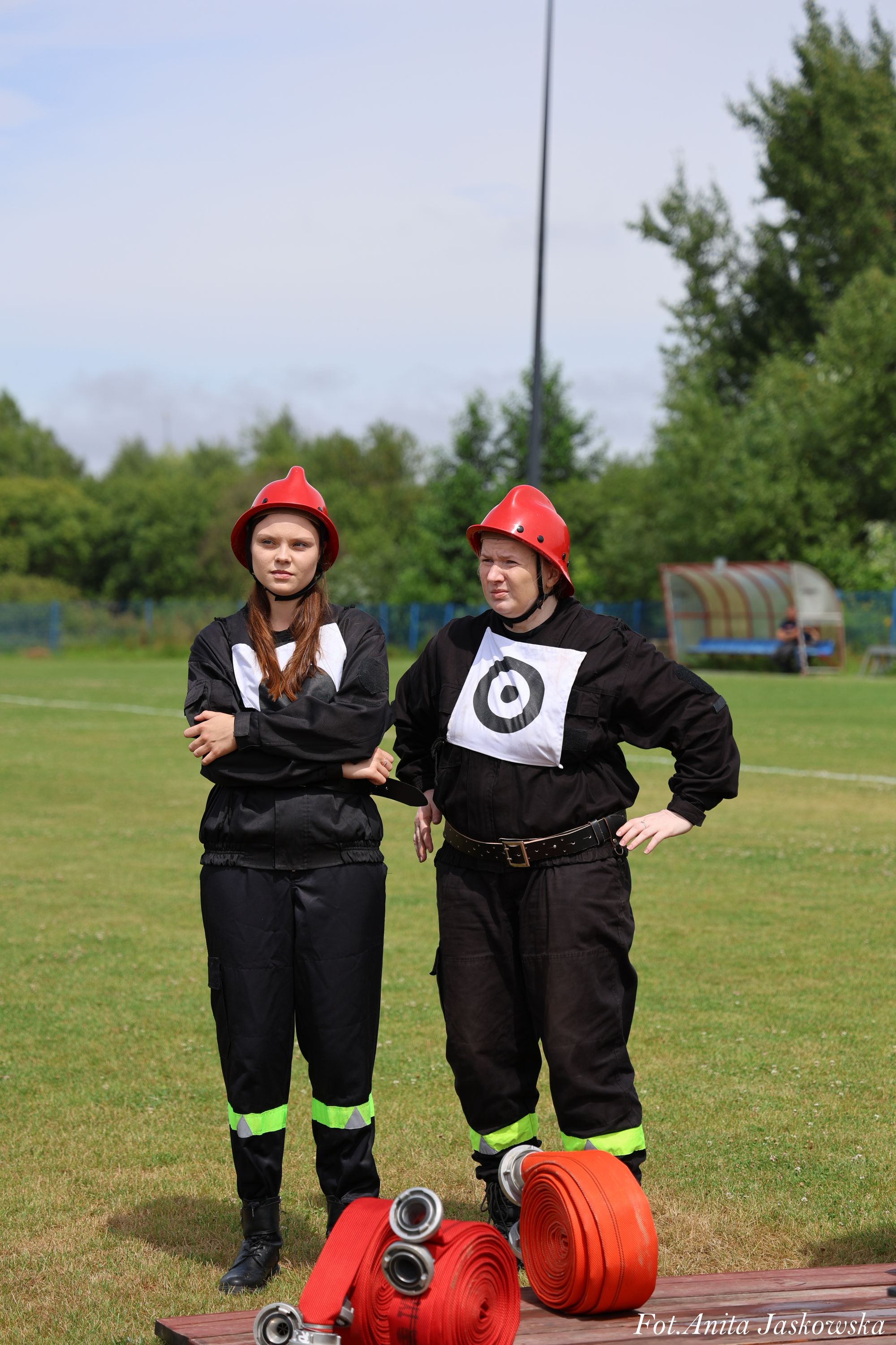Dwie osoby w czarnych ubraniach ze znakami na piersiach i czerwonych hełmach stoją na trawie, obok zwinięte czerwone węże strażackie.