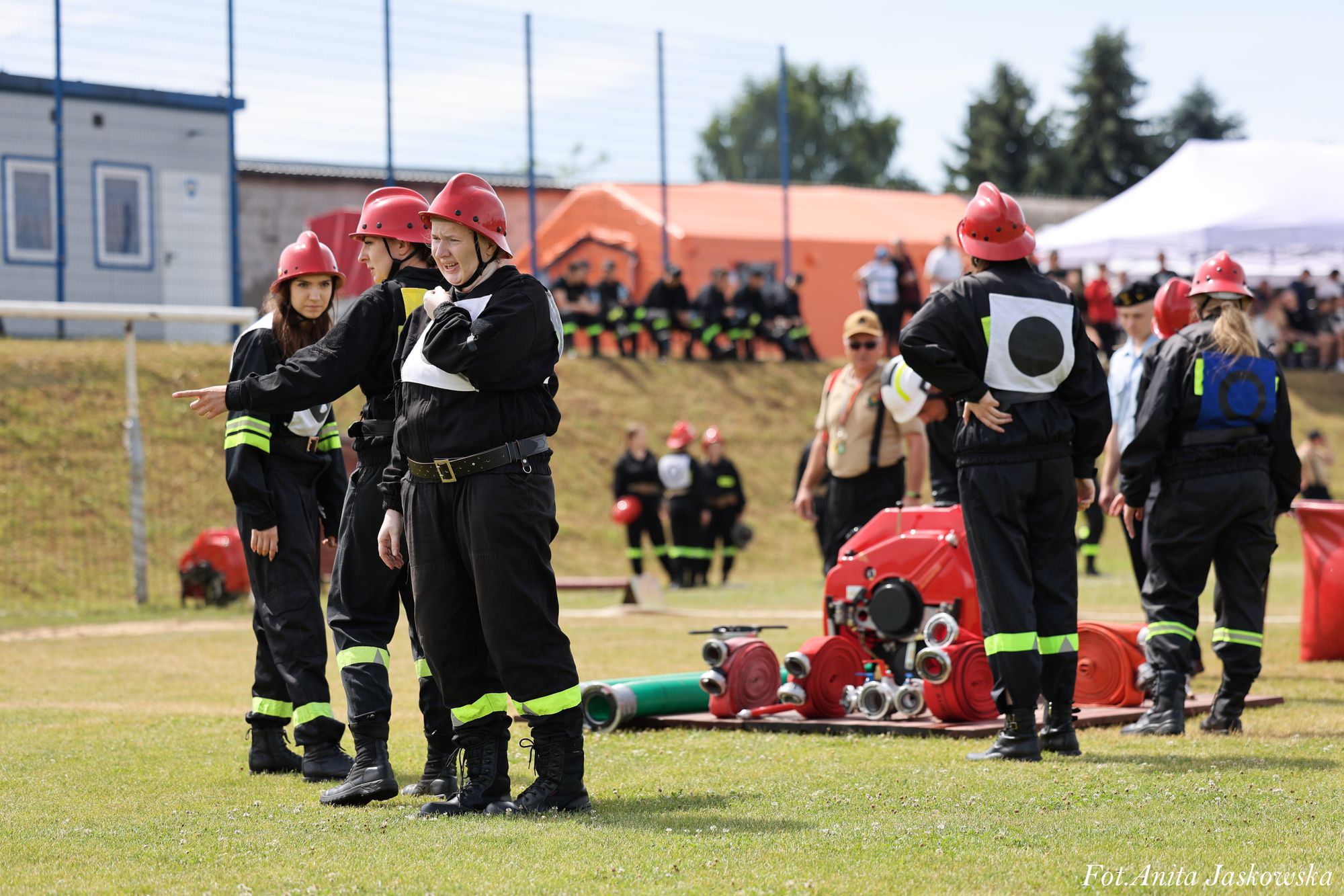 Grupa osób w czarnych mundurach z żółtymi pasami i czerwonych hełmach stoi na trawie obok sprzętu strażackiego pod gołym niebem podczas wydarzenia.
