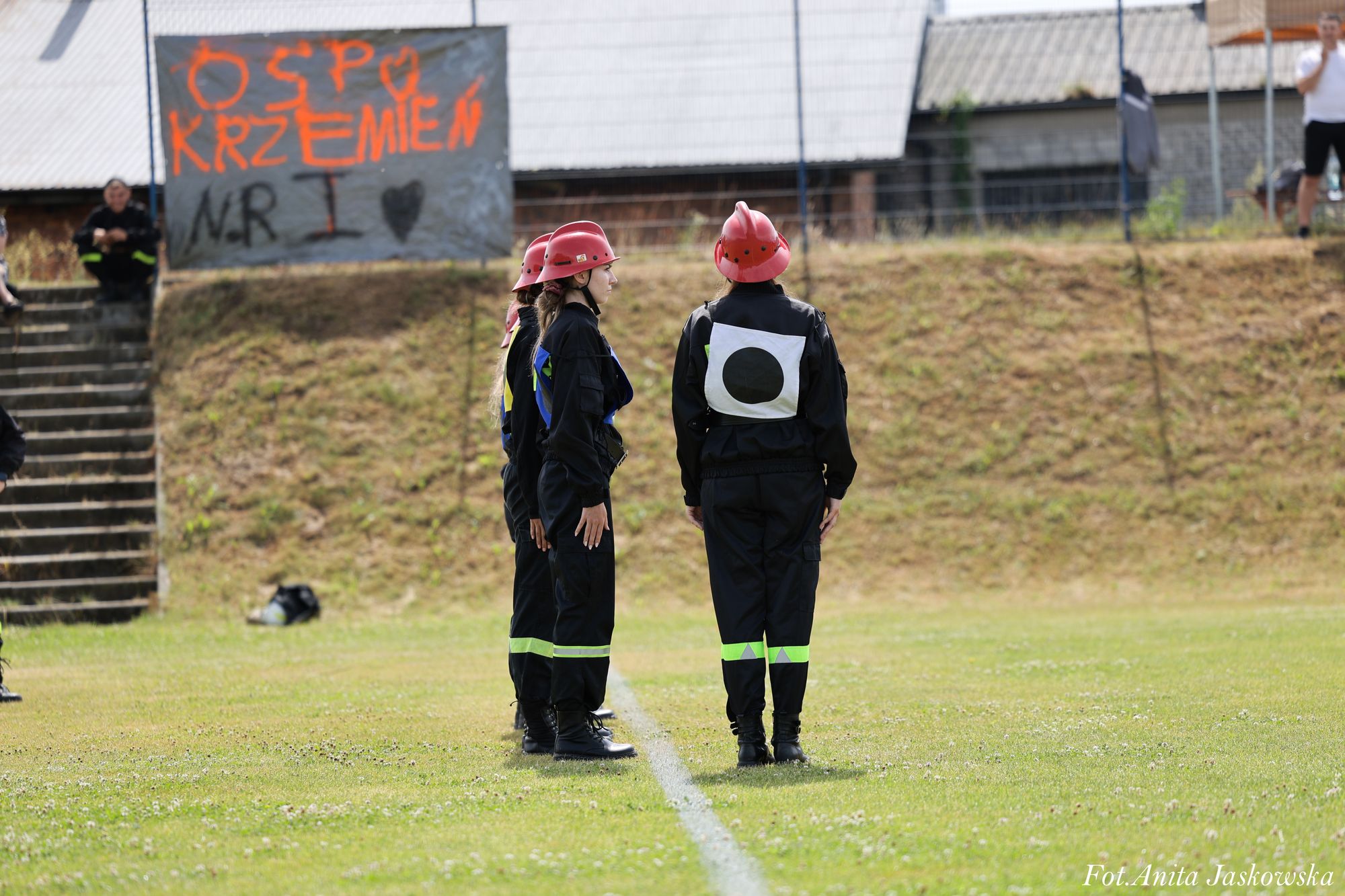 Trzy osoby w czarnych uniformach z czerwonymi hełmami stoją na zielonej trawie, za nimi wzgórze z napisem "OSP Krzemień" na szarym tle.