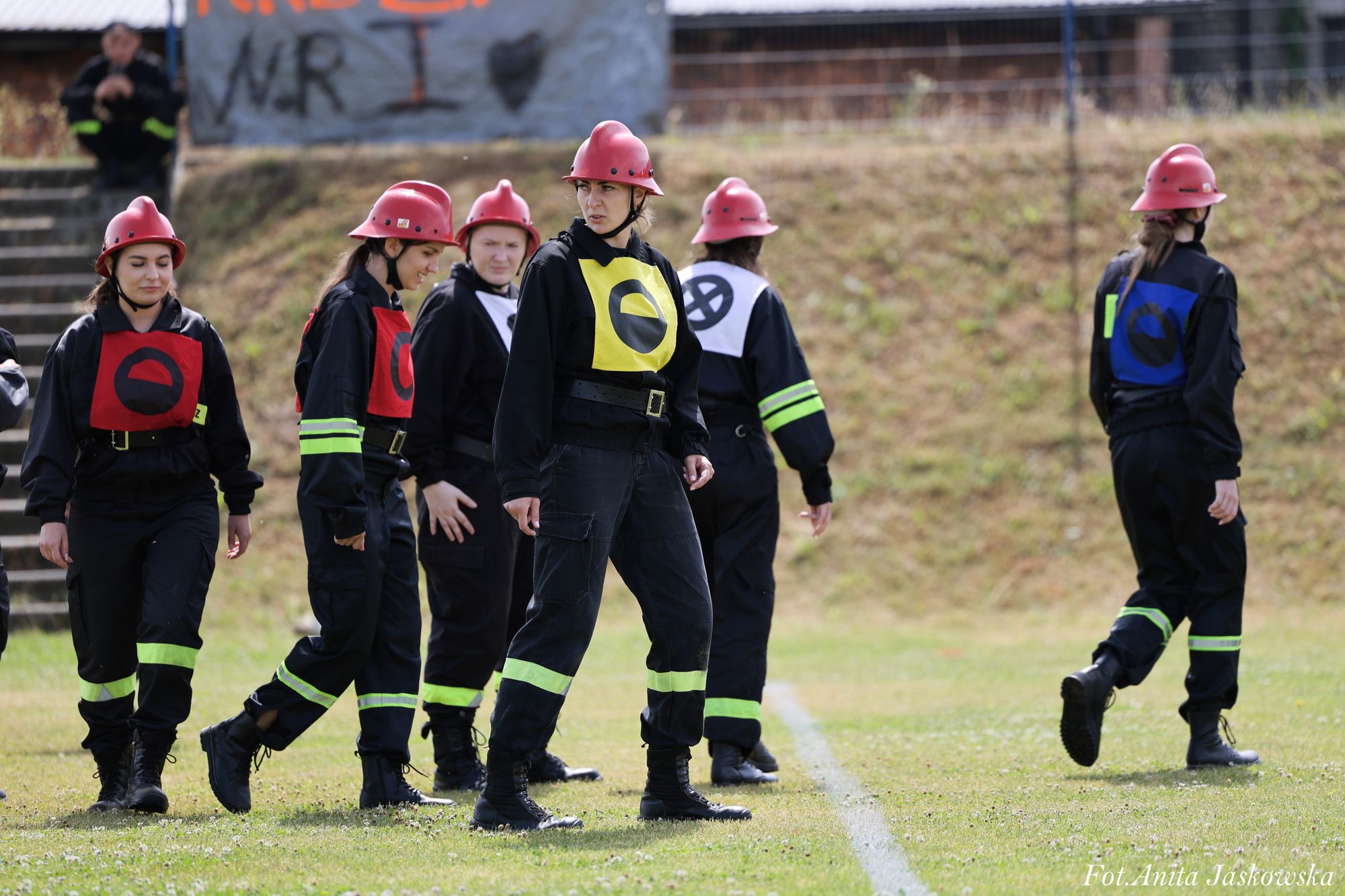 Grupa kobiet w czarnych kombinezonach z odblaskowymi paskami, czerwonych hełmach i kolorowych oznaczeniach na piersiach stoi i maszeruje po trawie na tle schodów i płotu.