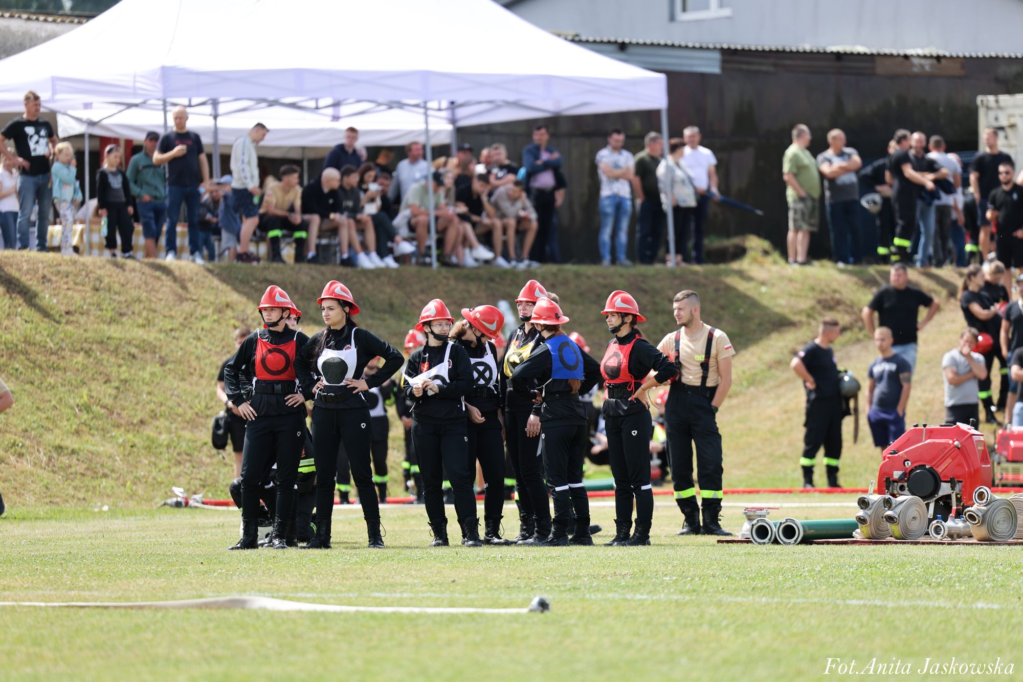 Grupa osób w czarnych ubraniach i czerwonych hełmach na trawie, z białymi i czerwonymi znakami na ubraniach, w tle ludzie pod białym namiotem i stojący na trawie.