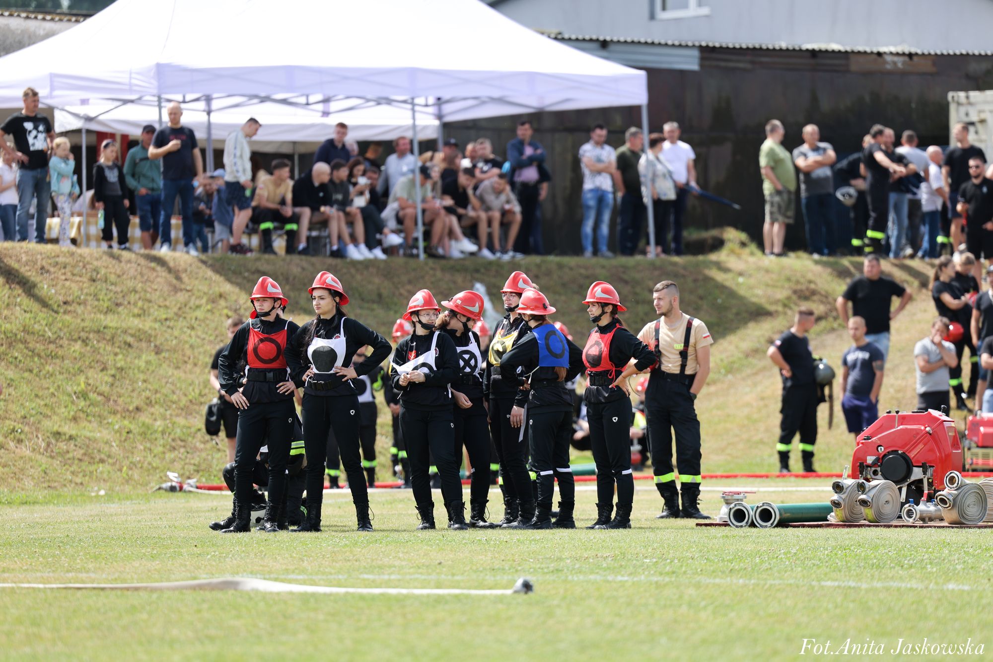 Grupa osób w czarnych mundurach i czerwonych hełmach stoi na zielonej trawie, w tle widoczne trybuny z ludźmi pod białym namiotem oraz sprzęt strażacki na trawie.