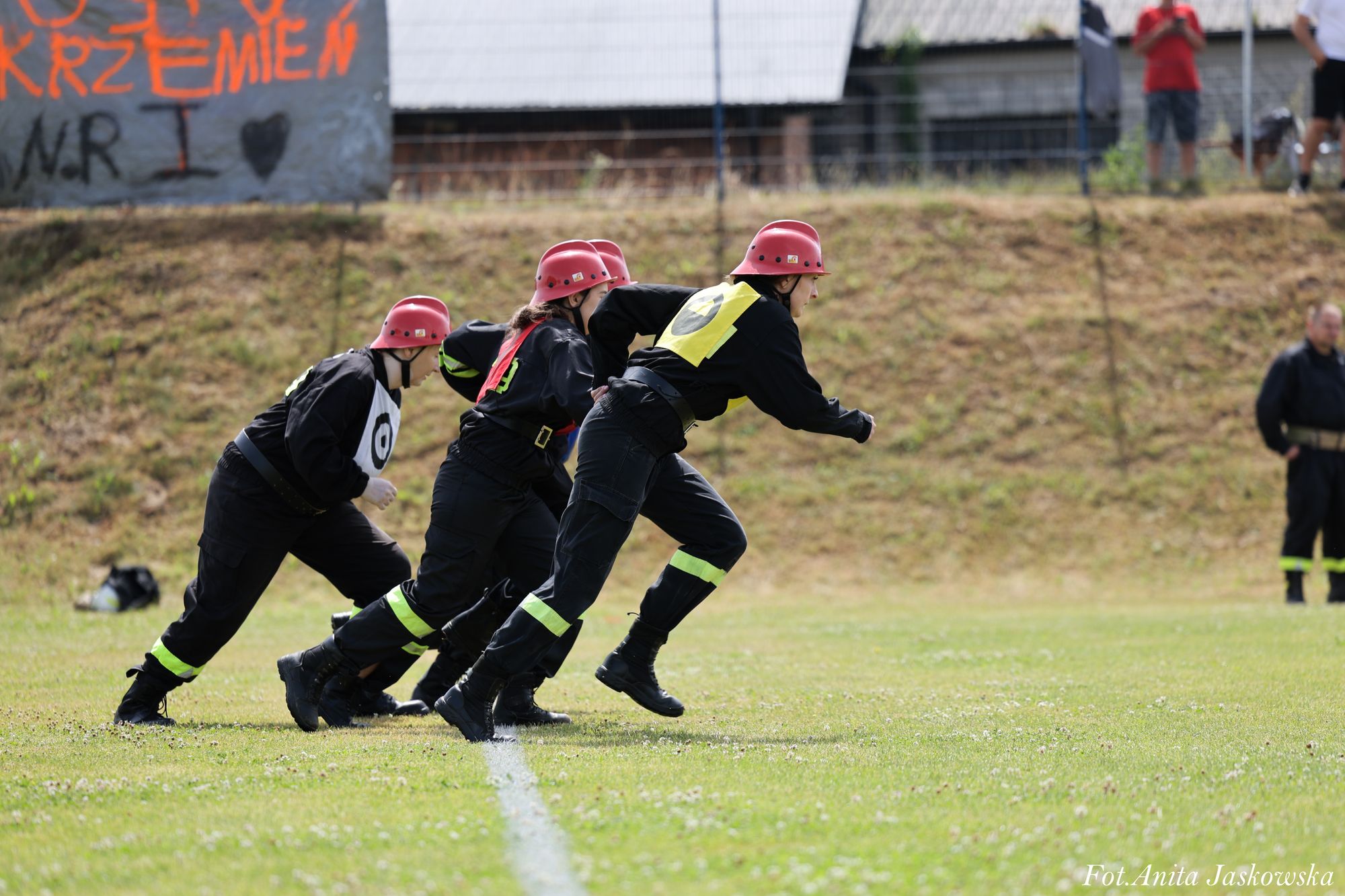 Cztery osoby w czarnych mundurach z żółtymi i białymi oznaczeniami oraz czerwonych hełmach rozpoczynają bieg na zielonym boisku. W tle trawa, płot i ludzie.
