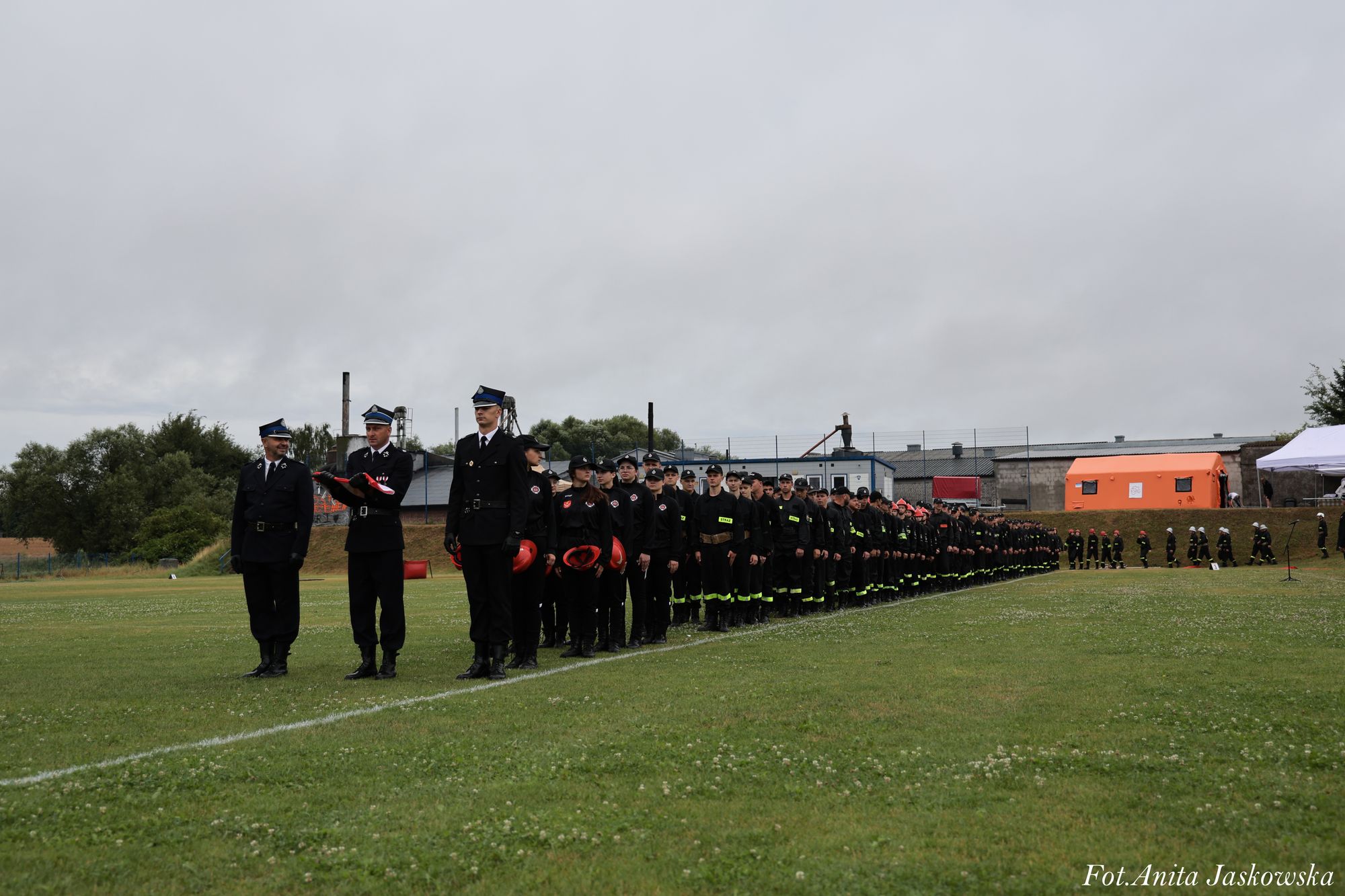 Ludzie w czarnych mundurach i hełmach ustawieni w długim szeregu na zielonym polu, za nimi budynki i szare, pochmurne niebo.