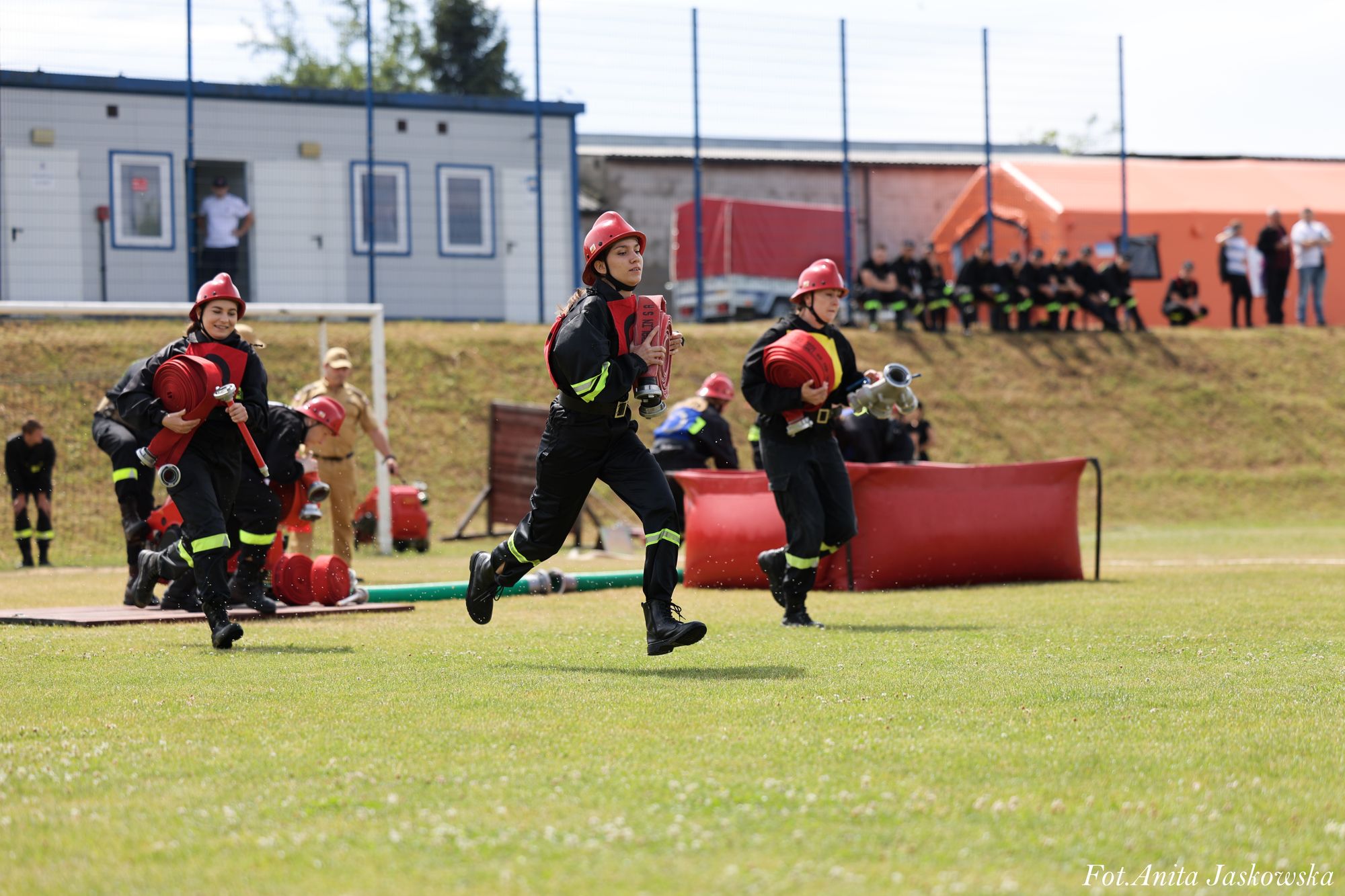 Trzy kobiety w czarnych mundurach i czerwonych hełmach biegną po zielonym boisku, każda niesie zwinięty czerwony wąż strażacki, w tle budynki i widzowie.