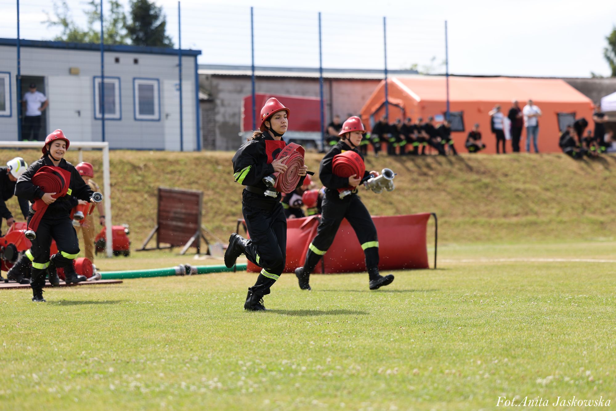 Trzy osoby w czarnych strojach strażackich z czerwonymi hełmami biegną po zielonym boisku trzymając zwinięte czerwone węże strażackie, w tle ludzie i pomarańczowy namiot.