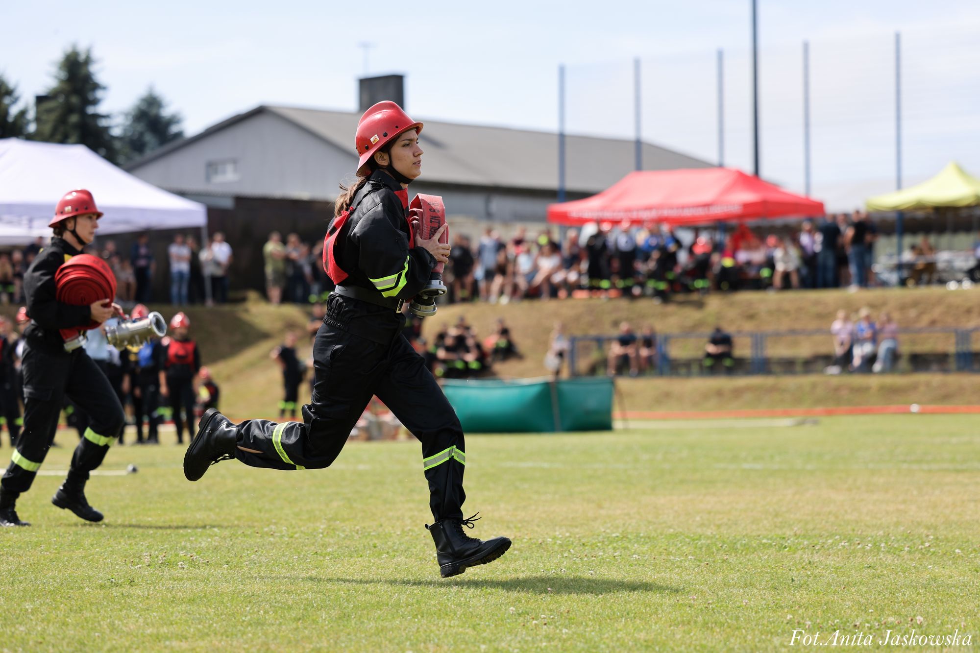 Dwie osoby w czarnych mundurach i czerwonych hełmach strażackich biegną po zielonym boisku, trzymając zwinięte czerwone węże strażackie, w tle rozmyta widownia i namioty.