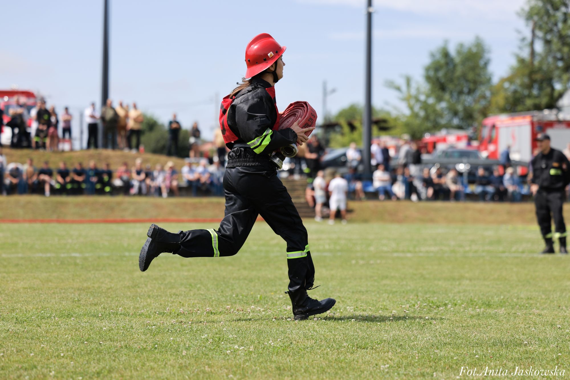 Strażak w czarnym uniformie z żółtymi pasami i czerwonym hełmem biegnie po zielonej trawie, trzymając zwinięty czerwony wąż strażacki, w tle rozmazani ludzie i wozy strażackie.