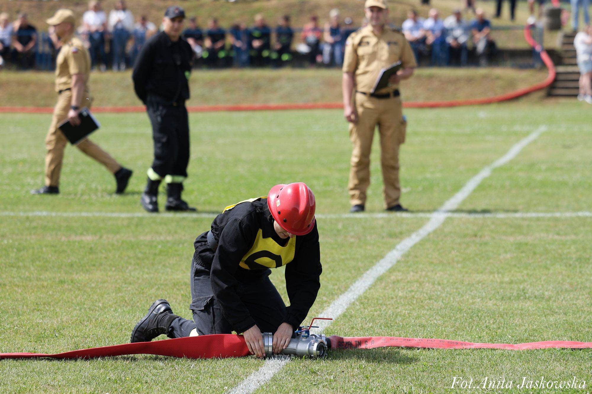 Osoba w czerwonym kasku i czarnym ubraniu z żółtym numerem na kolanach na zielonej trawie rozłącza czerwony wąż strażacki, w tle trzy osoby w mundurach i rozmyty tłum.