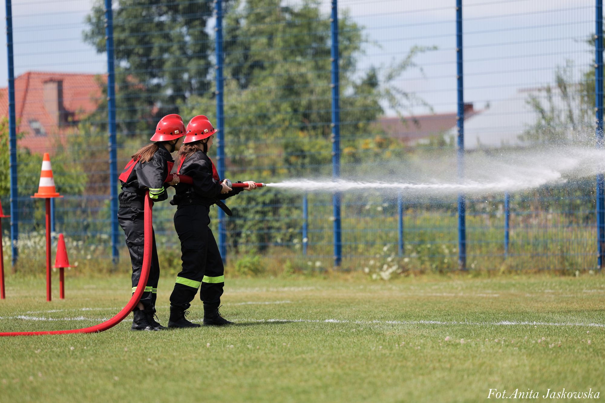 Dwie osoby w czarnych strojach strażackich i czerwonych hełmach trzymają wąż strażacki, z którego leci strumień wody, stojąc na zielonej trawie na tle niebieskiego ogrodzenia.