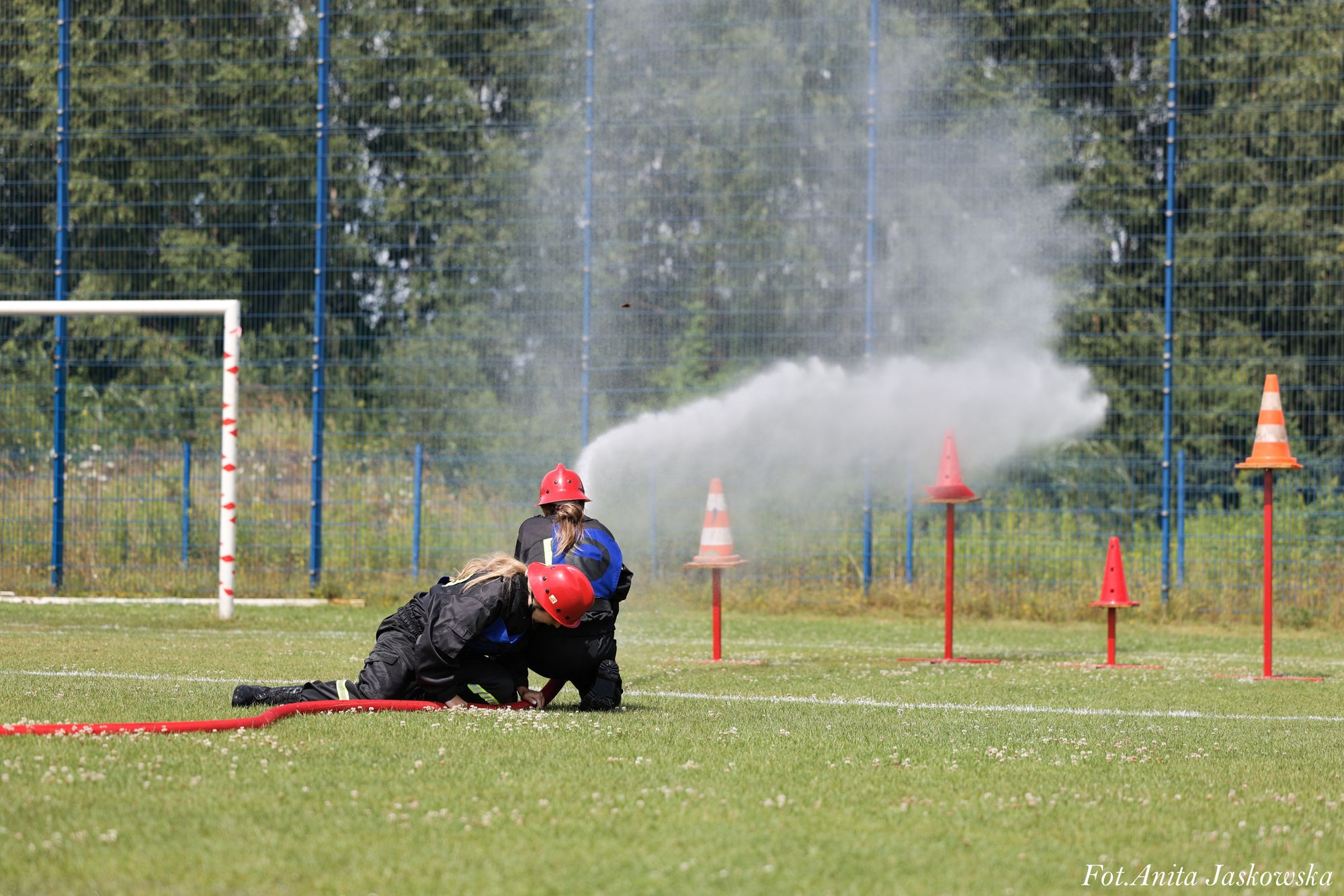 Dwie osoby w czarnych kombinezonach i czerwonych hełmach strumieniem wody z węża gaszą pomarańczowe pachołki na zielonym boisku sportowym.