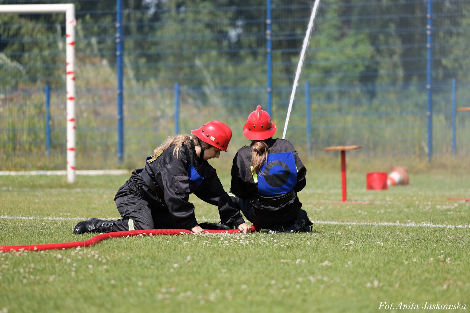 Dwie osoby w czarnych ubraniach i czerwonych hełmach na zielonej trawie, trzymające czerwony wąż strażacki, w tle ogrodzenie i biało-czerwona bramka.