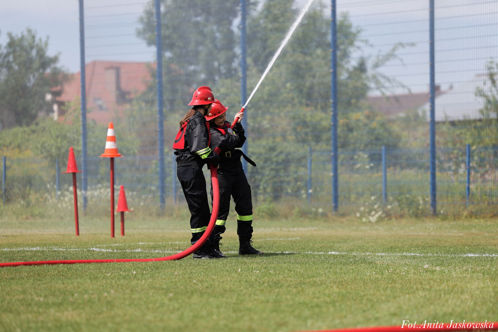 Dwie osoby w czarnych kombinezonach i czerwonych hełmach trzymają czerwoną wąż strażacki, z którego leci strumień wody na zielonym boisku.
