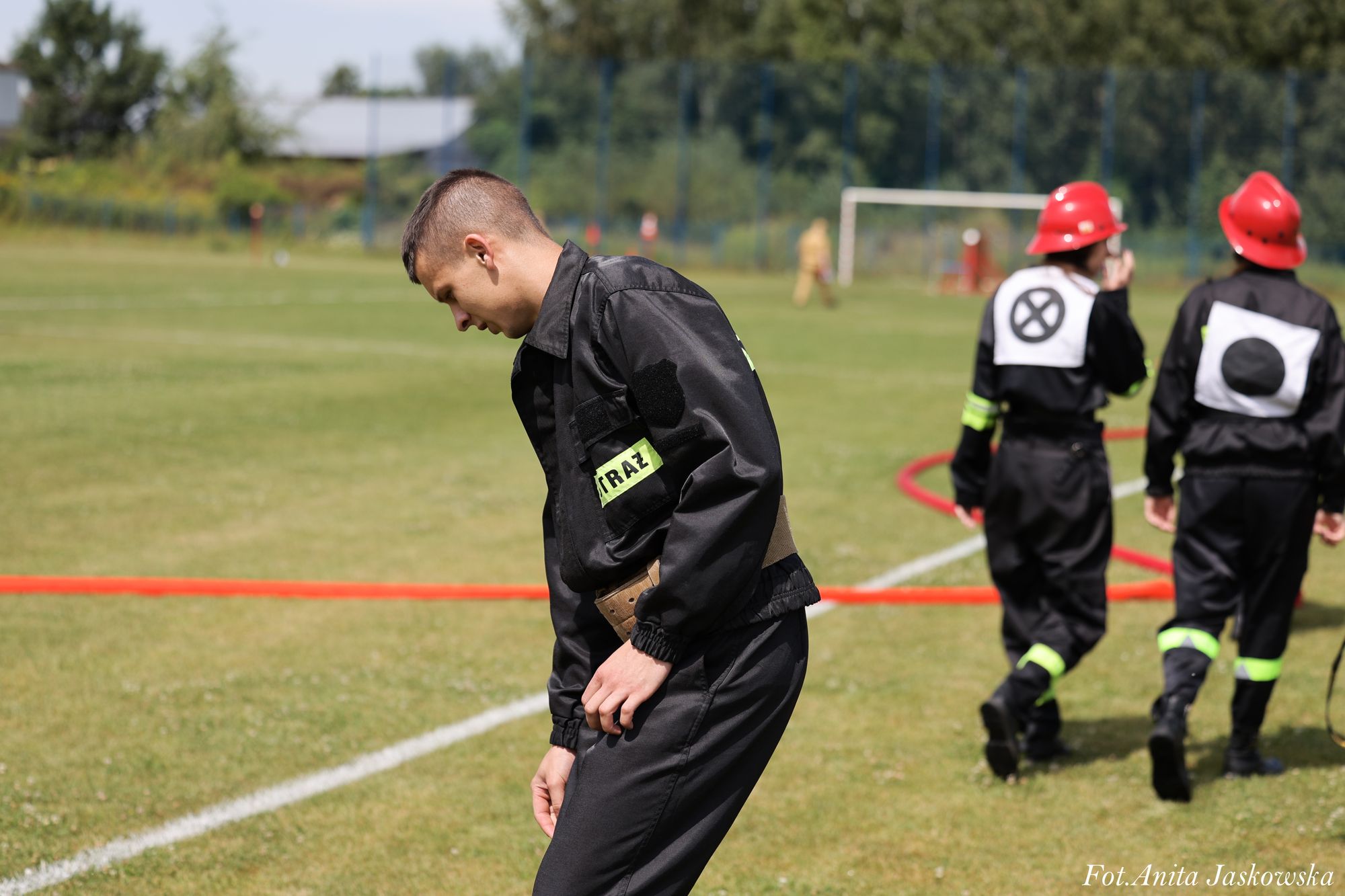 Młody mężczyzna w czarnym stroju strażackim z żółtą naszywką "STRAŻ" pochylony na trawie, w tle dwie osoby w czarnych ubraniach i czerwonych hełmach.