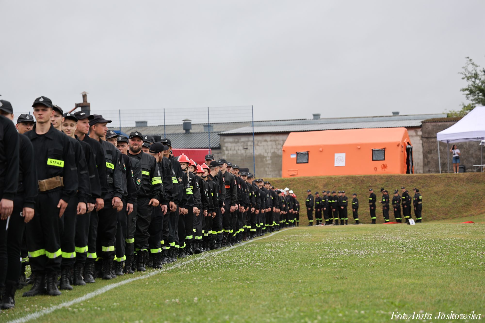 Szereg osób w czarnych mundurach z żółtymi odblaskowymi pasami stoi na linii białej na zielonej trawie, w tle namiot pomarańczowy i biały oraz budynki.