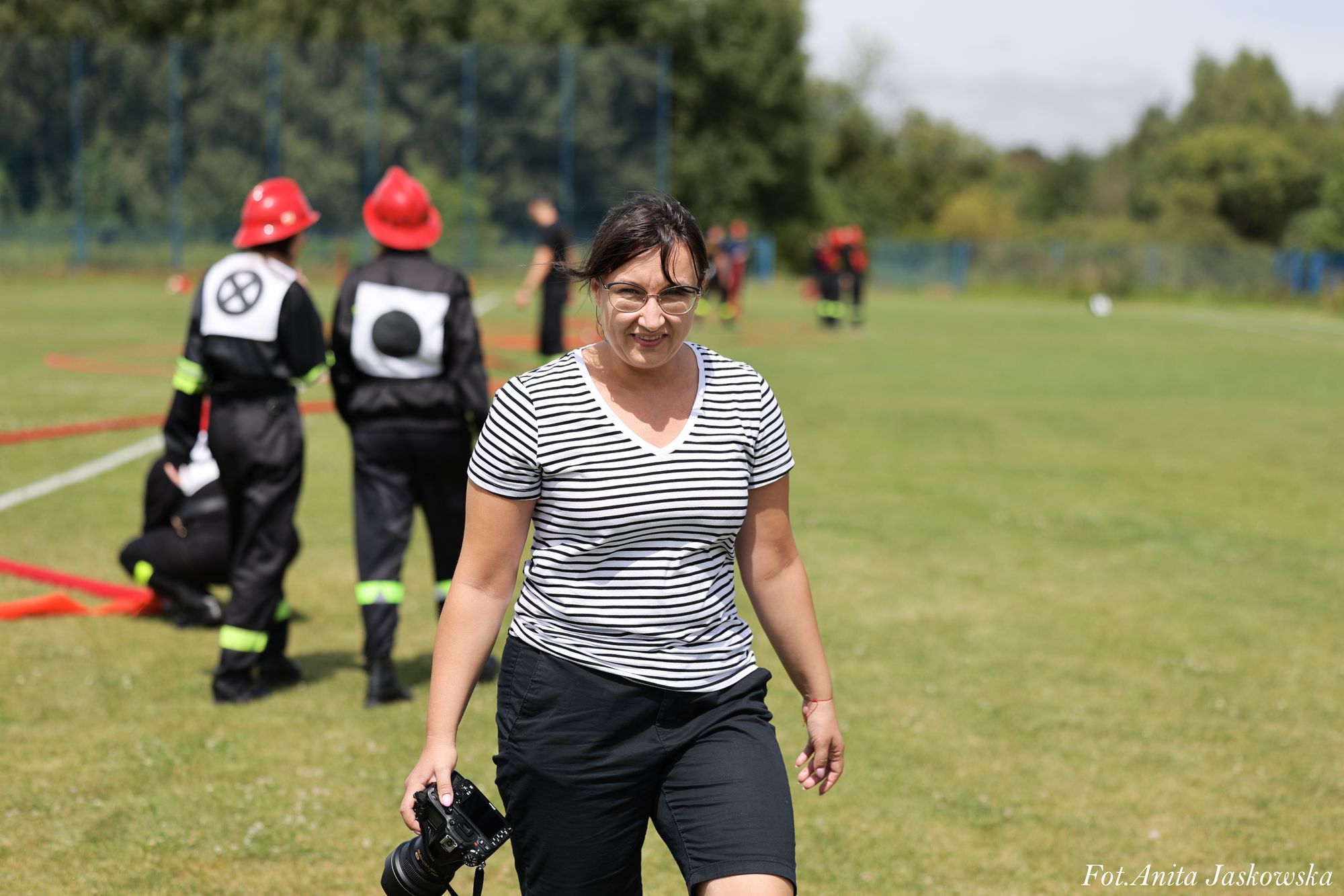 Kobieta w okularach, w białej koszulce w czarne paski i czarnych spodniach, trzyma aparat fotograficzny na tle zielonej trawy i osób w strojach strażackich.