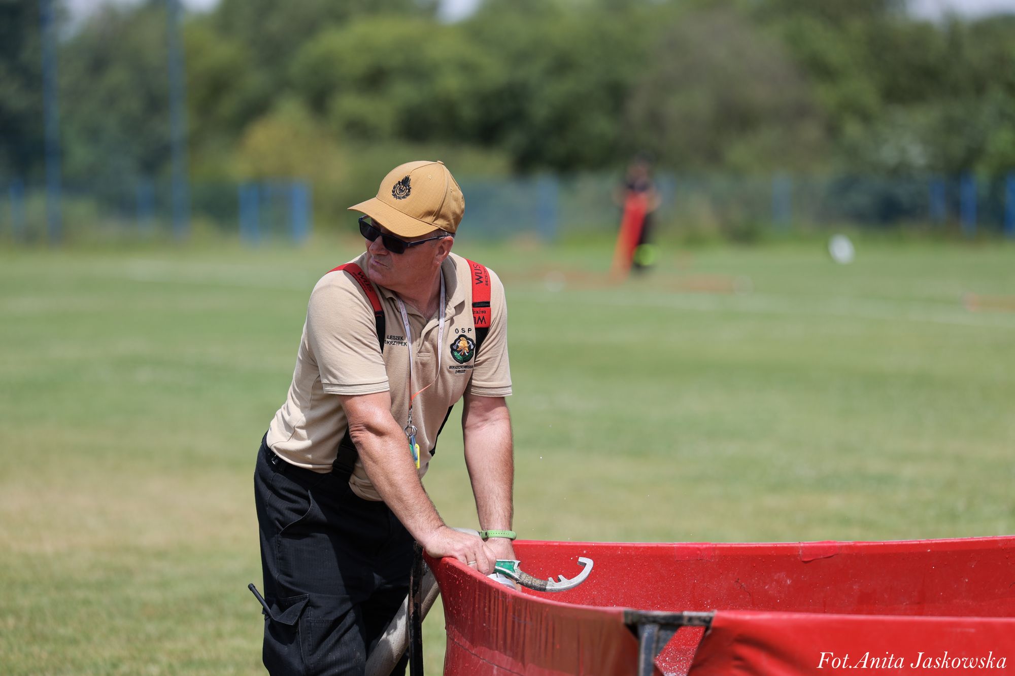 Mężczyzna w beżowej koszulce, czarnych spodniach i beżowej czapce pochyla się nad czerwonym zbiornikiem na trawiastym boisku.
