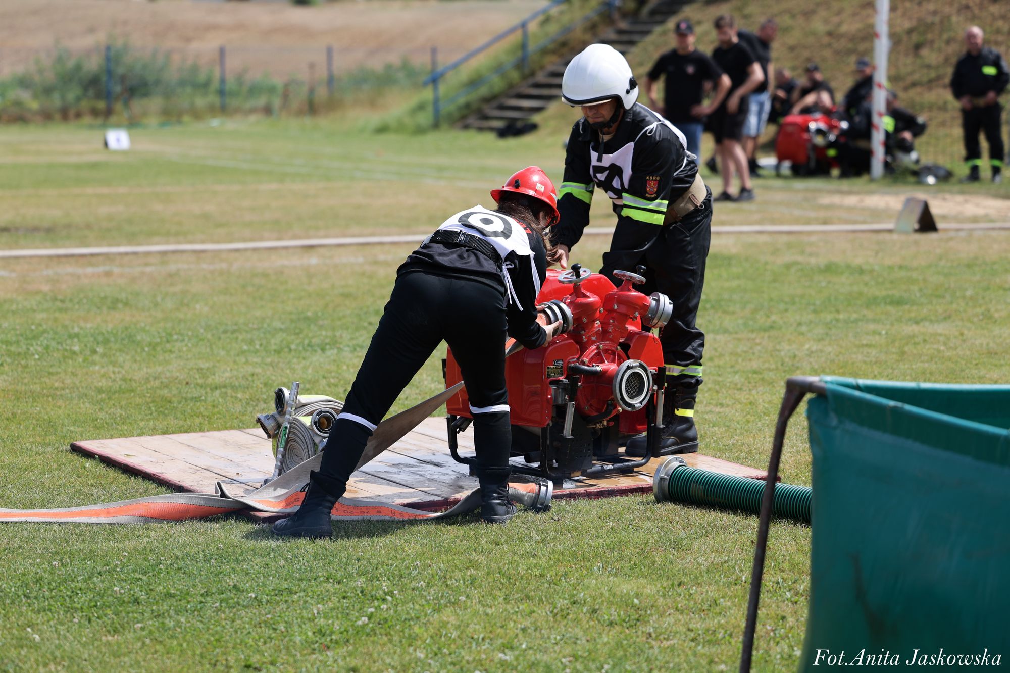Dwóch strażaków w czarnych mundurach i hełmach (biały i czerwony) podłącza wąż do czerwonej pompy na zielonej trawie na boisku sportowym.