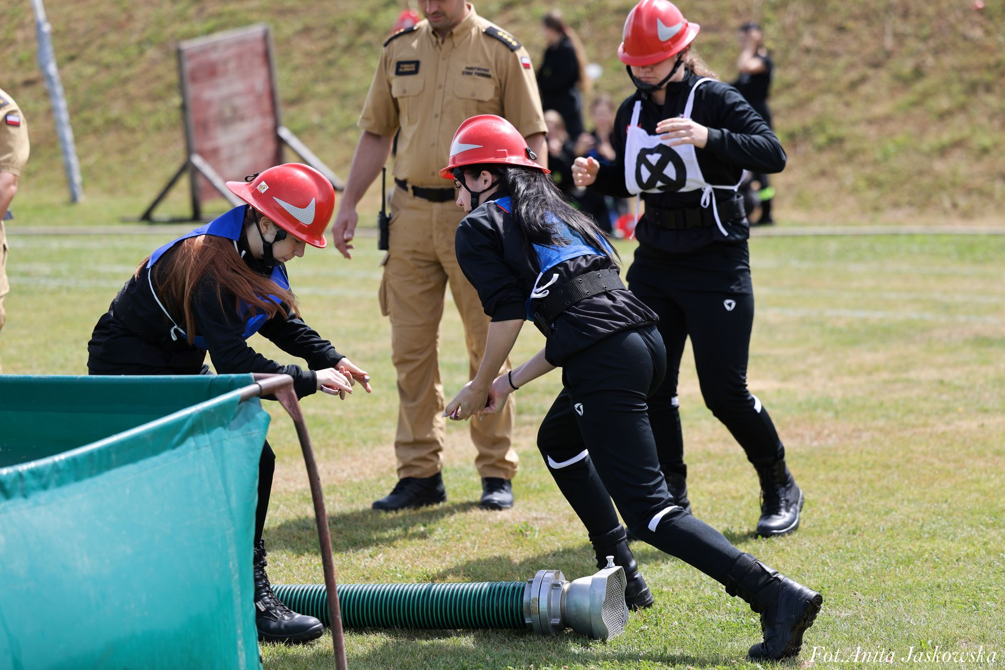 Trzy osoby w czarnych ubraniach i czerwonych kaskach manipulują zielonym wężem strażackim na trawniku, obok stoi mężczyzna w beżowym mundurze.