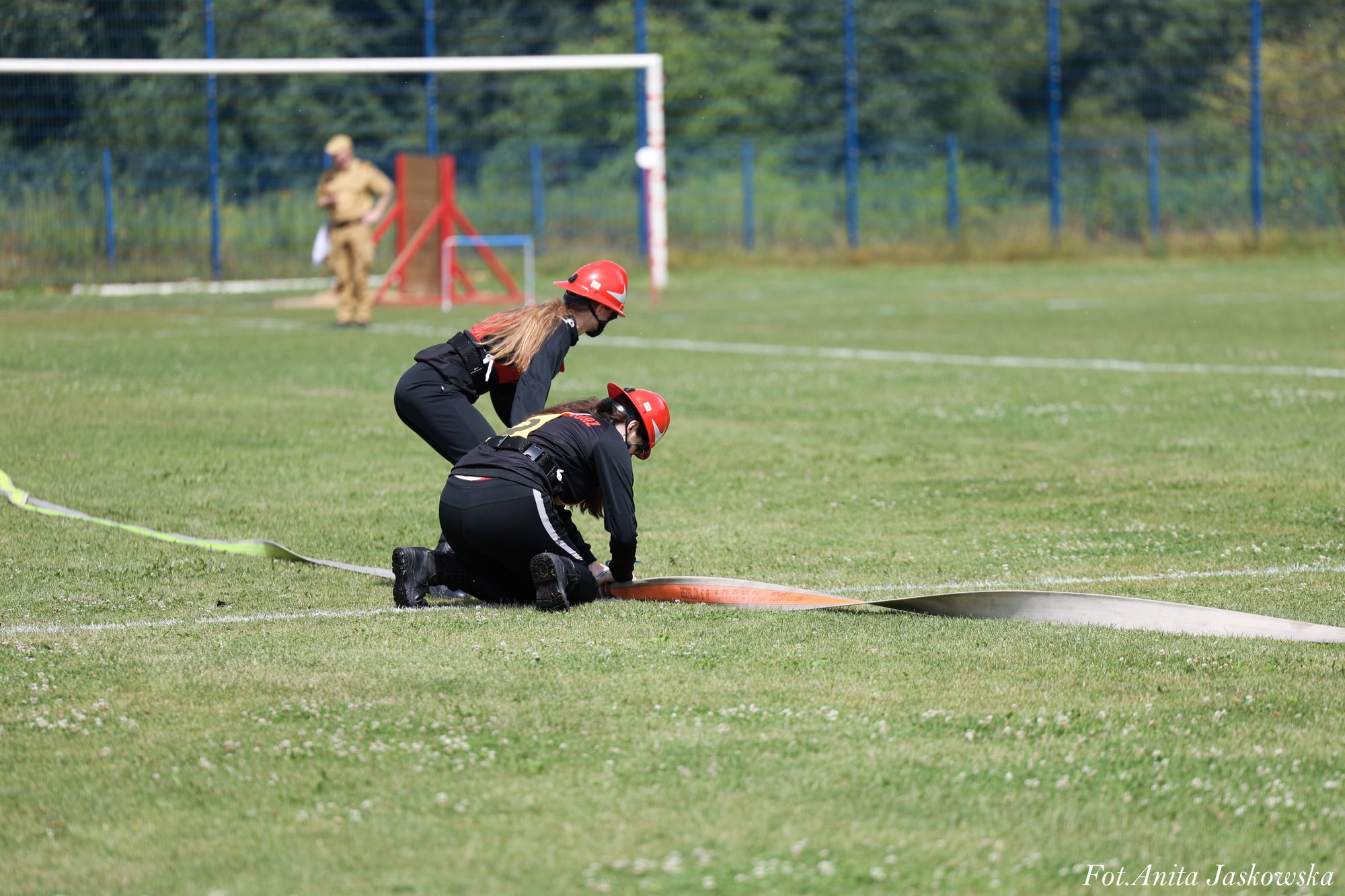 Dwie osoby w czarnych ubraniach i czerwonych hełmach klęczą na zielonej trawie, rozwijając pomarańczowy i szary wąż strażacki, w tle bramka i ogrodzenie.