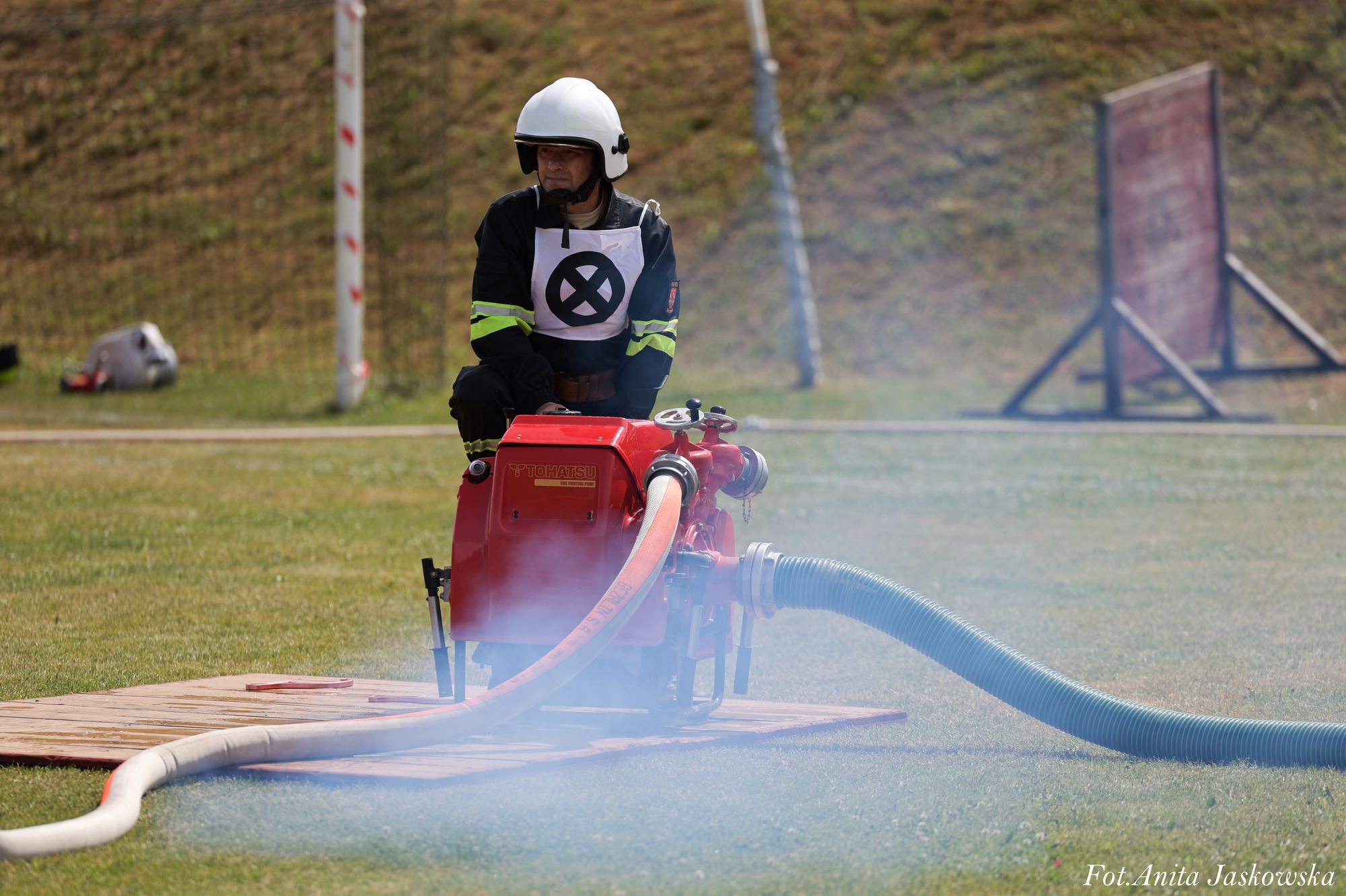 Mężczyzna w białym kasku i czarnym ubraniu z odblaskowymi paskami siedzi przy czerwonej pompie pożarniczej na trawie, wokół unosi się lekka mgła lub dym.