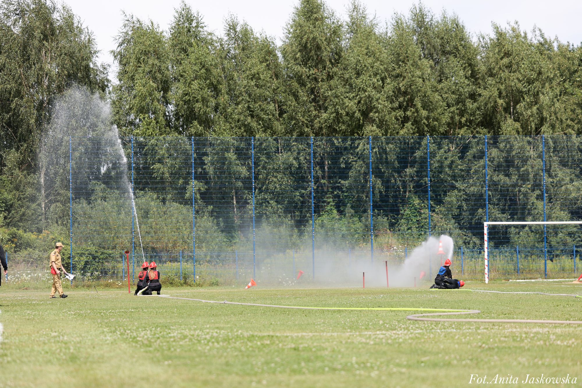 Cztery osoby w ubraniach strażackich i czerwonych hełmach na zielonym boisku, dwie z nich podlewają wodą z węży, w tle wysoka siatka i drzewa.