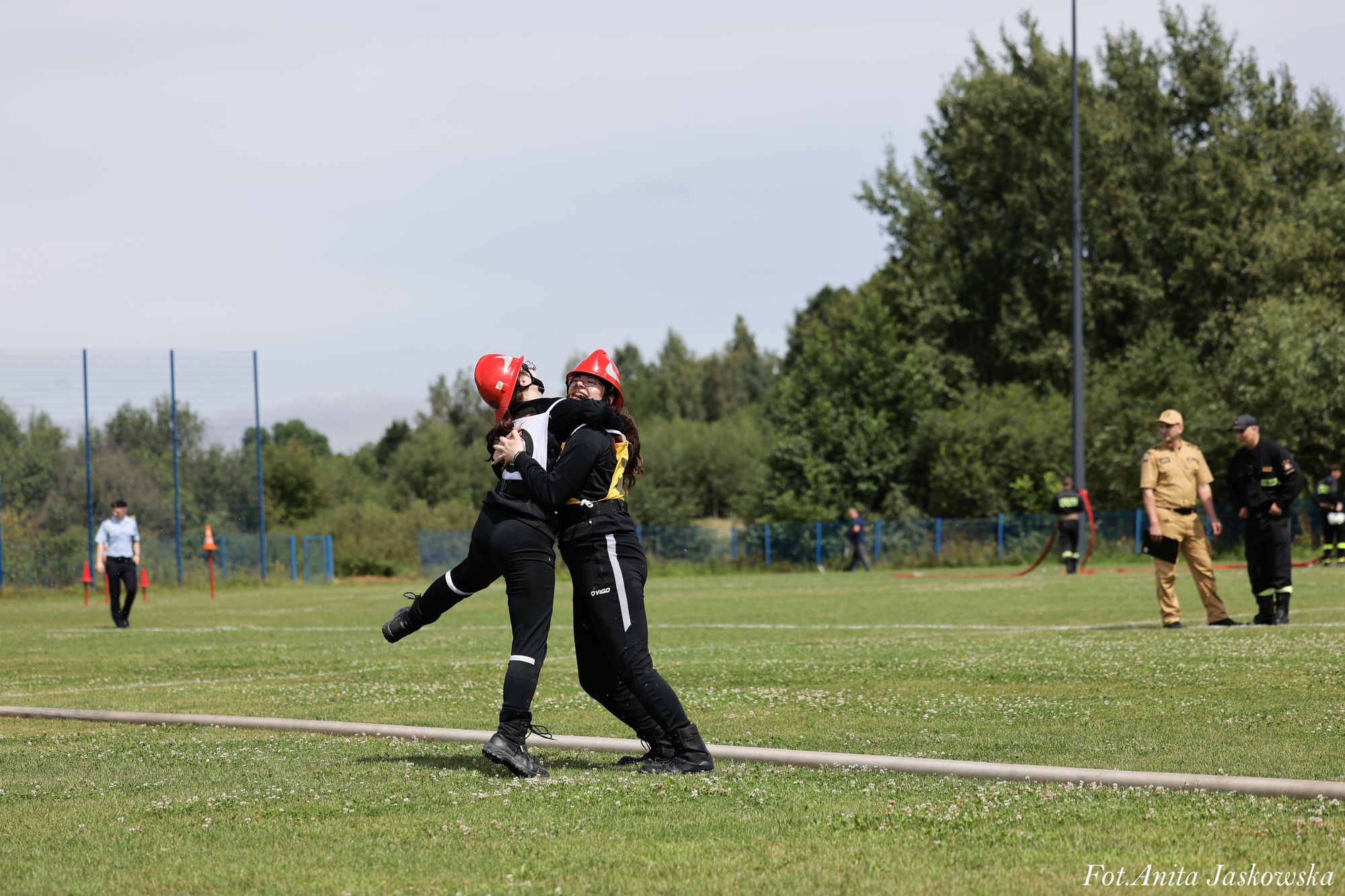 Dwie osoby w czarnych strojach i czerwonych hełmach na zielonej trawie, jedna podnosi drugą, w tle drzewa, ludzie w mundurach, niebieskie ogrodzenie.