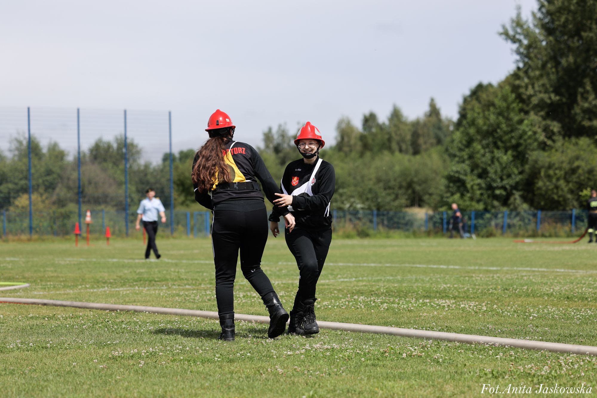 Dwie osoby w czerwonych kaskach i czarnych ubraniach stojące na zielonym boisku, w tle drzewa i płot, jedna osoba idąca w oddali.