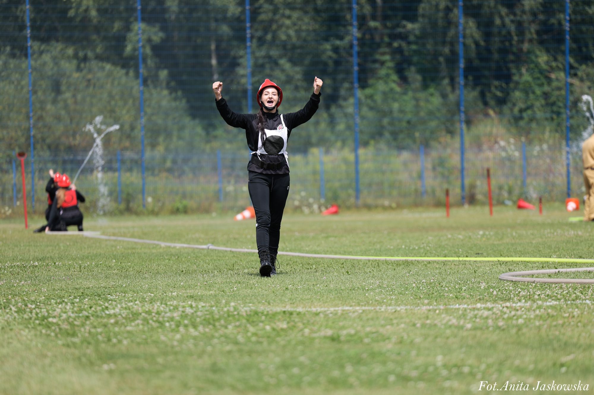 Kobieta w czerwonym hełmie i czarnym ubraniu z białym pasem, stoi na zielonym boisku trawiastym, w tle siatka i dwie osoby w czerwonych hełmach oraz woda tryskająca z węża.